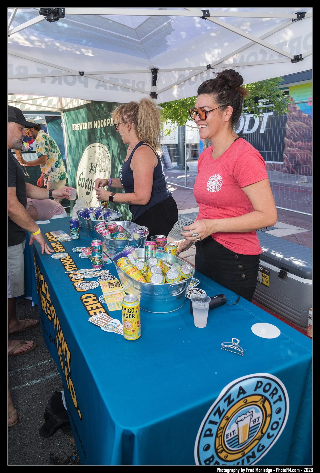 People at a booth with a blue tablecloth displaying the logo of Pizza Port Brewing Co., selling canned drinks including beer and soda at an outdoor event under a white canopy.
