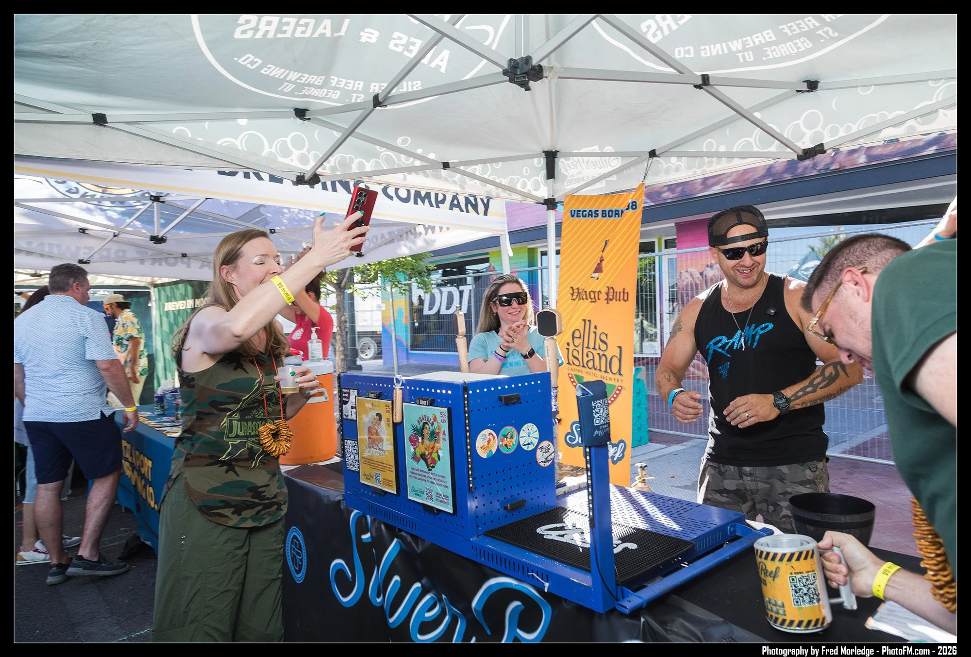 People gather at an outdoor booth with a blue sign that reads 'Slovers.' A woman in a camouflage shirt takes a photo with her phone, while a man in sunglasses and a black tank top smiles nearby. Other people are in the background, some wearing wristb