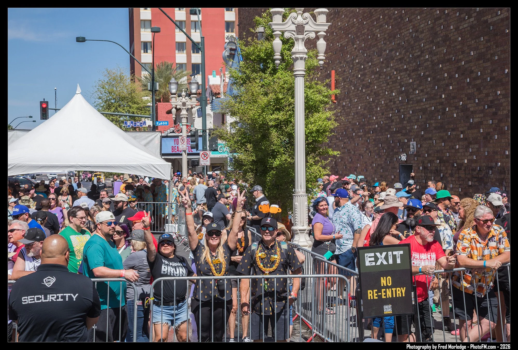 Crowd of people standing behind security barriers at an outdoor event, with some making peace signs and talking. Security personnel are present, and a tent is set up nearby. The background includes buildings, trees, streetlights, and a large brick wa