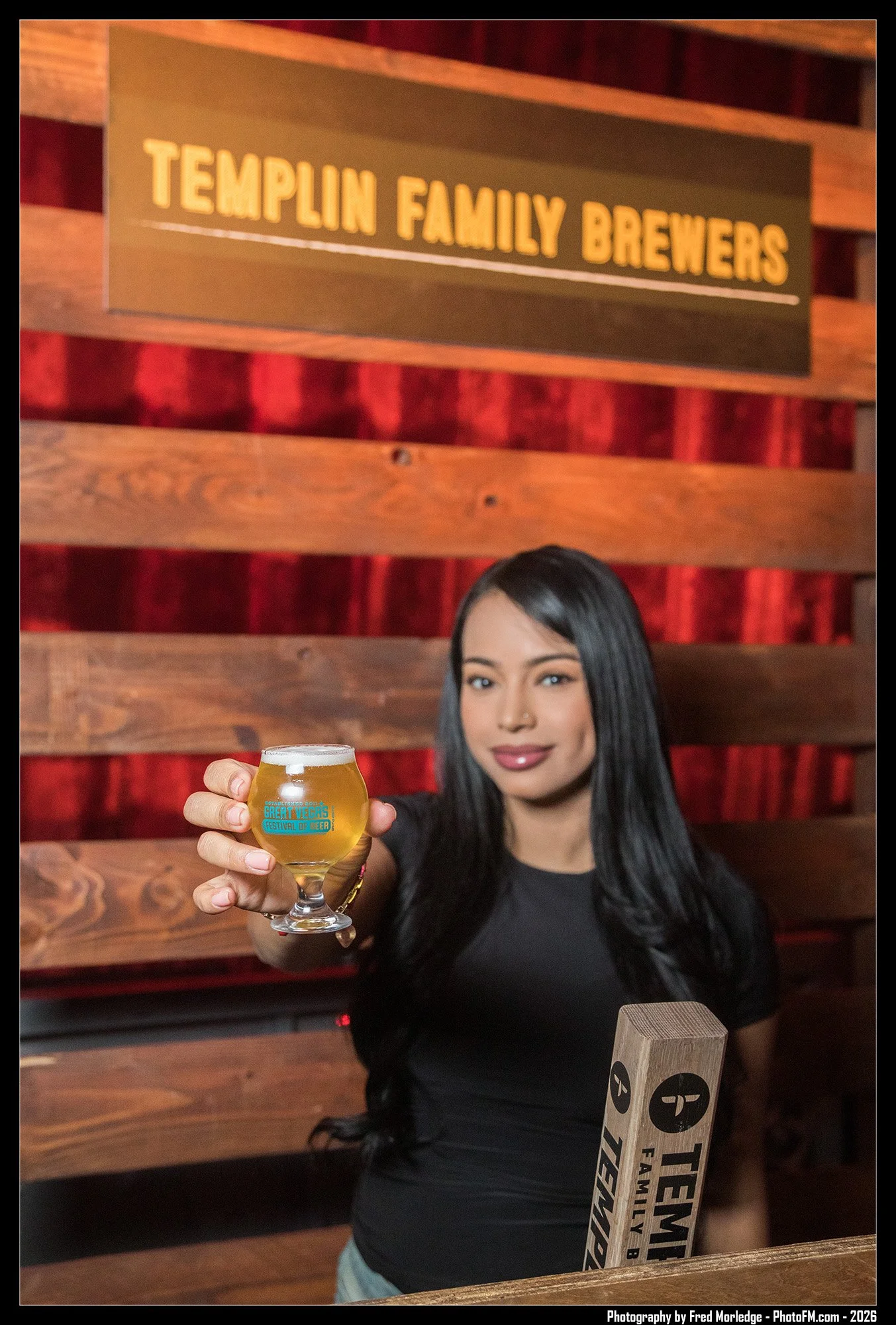 Woman with long black hair smiling and holding a glass of beer at a brewery. She is wearing a black shirt and standing in front of a wooden wall with a sign that reads 'Templin Family Brewers'.