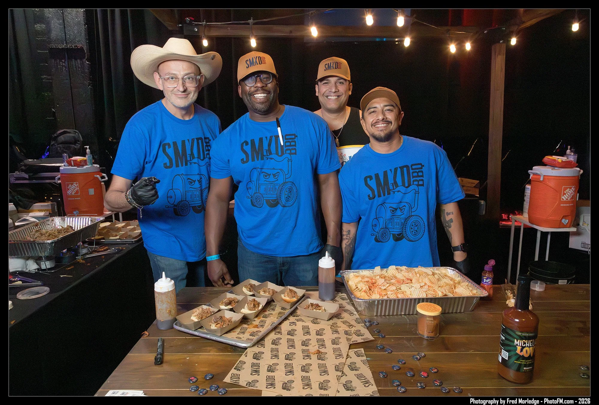 Four men smiling at the camera behind a table with food at a barbecue event. They are wearing blue "SMKYD BBQ" T-shirts, with one wearing a large cowboy hat and another wearing baseball caps. The table has paper food trays, bottles of barbecue sauce,