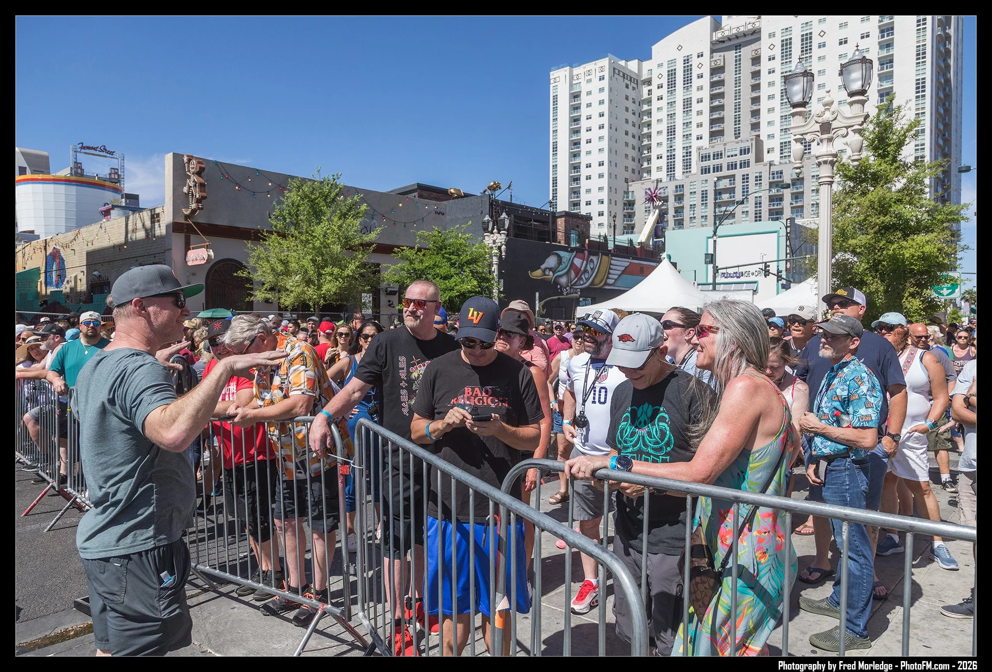 Crowd of people behind metal barricades at an outdoor event, with some individuals using mobile phones, under a clear blue sky with city buildings and trees in the background.