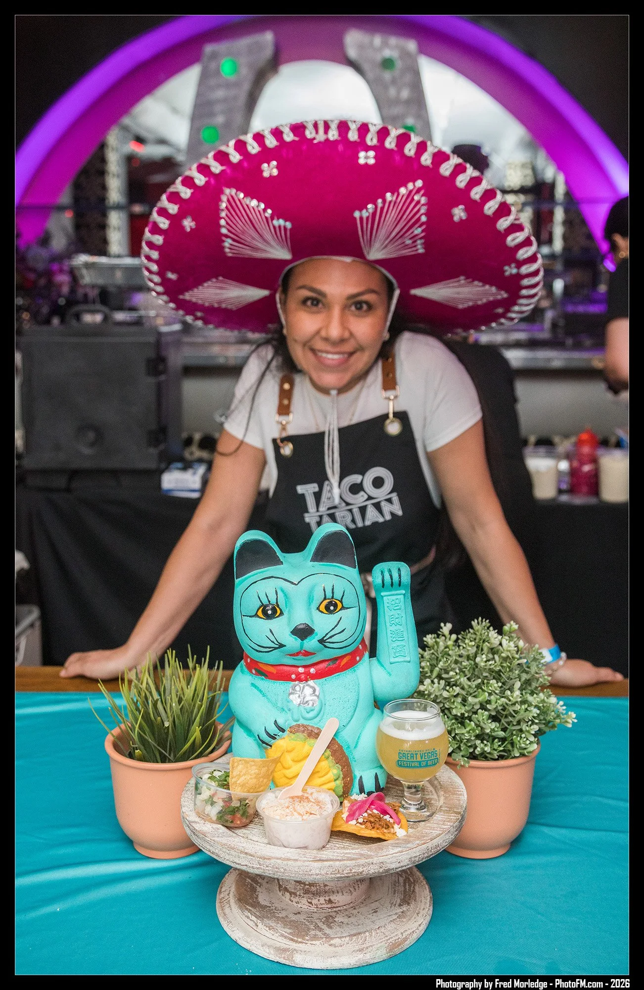 A woman wearing a large pink sombrero hat stands behind a table decorated with a Día de los Muertos themed display. The display includes a turquoise ceramic cat figurine, plants, and a small food plate with tacos, chips, and desserts. There is also a