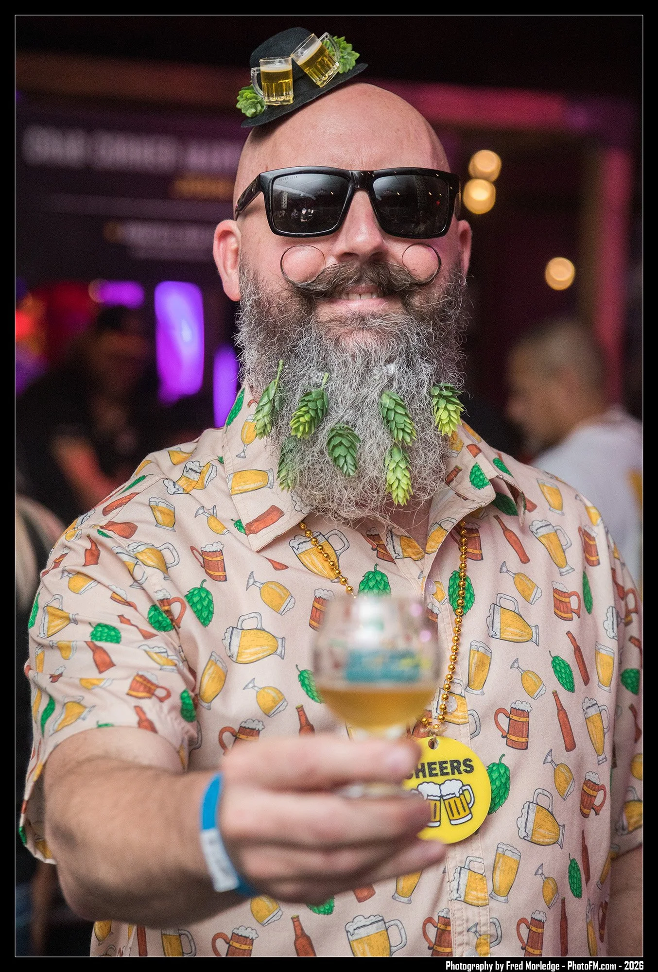 A man with a bald head, a gray beard, and a handlebar mustache is smiling and wearing large black sunglasses. He has a small black hat with miniature beer mugs and green hops decorations on top. He is dressed in a light-colored shirt with a beer mug 