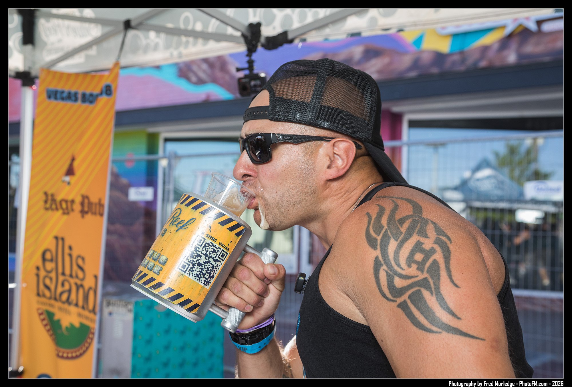 A muscular man wearing a black tank top, black sunglasses, and a black mesh cap is drinking from a large beer mug outdoors. The mug has a yellow label with QR code and text. In the background, there are colorful banners and a canopy with a scenic mou