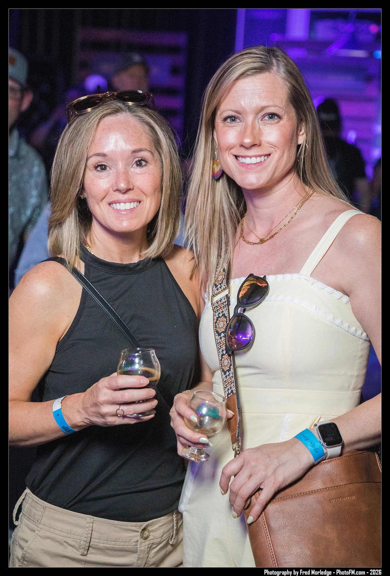 Two women smiling at a social event, each holding a glass of wine, wearing casual summer clothes, with purple lighting in the background.