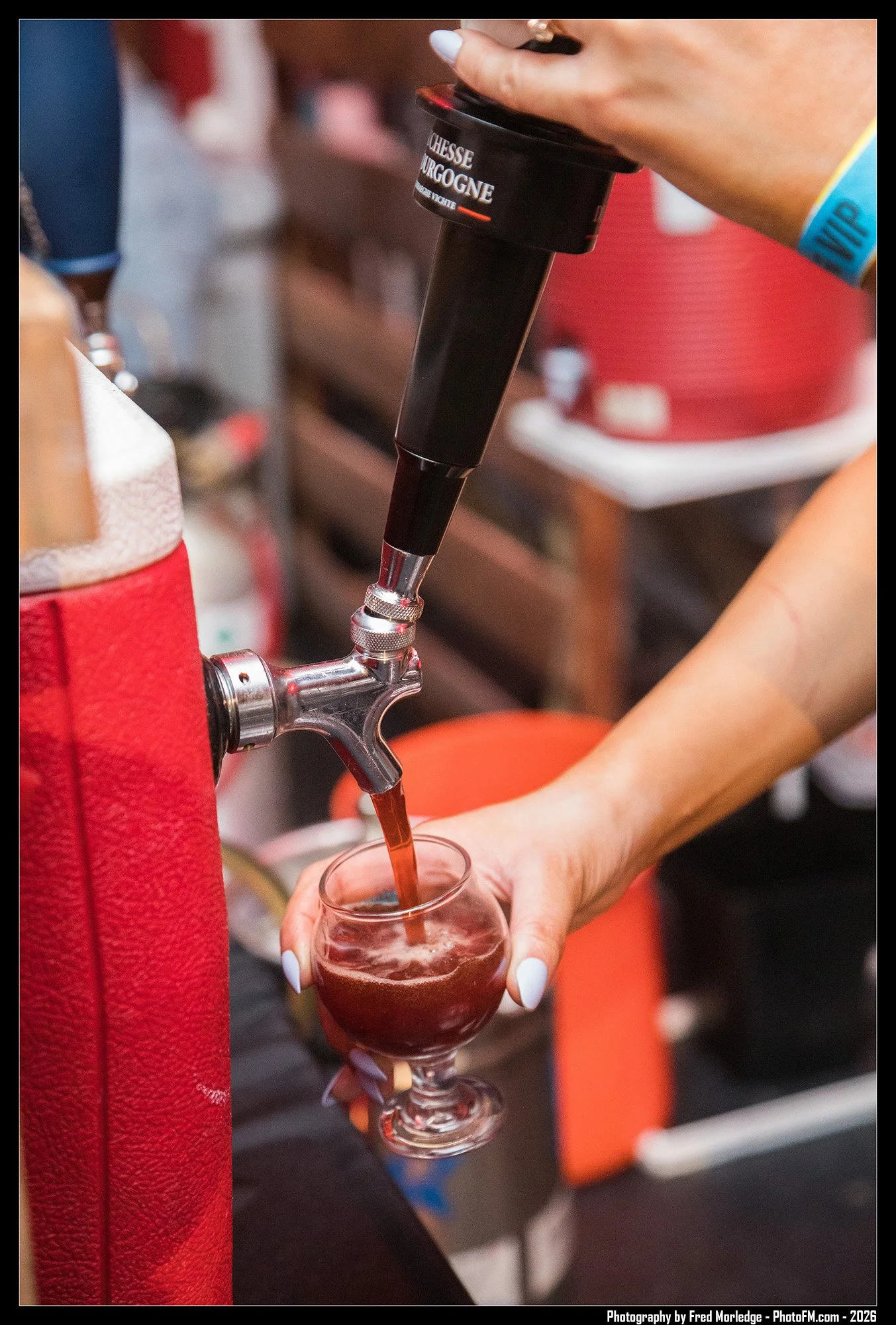 A person is holding a glass under a tap pouring a dark beverage, possibly red wine or juice. The tap is attached to a dispenser labeled "FROMAGE" and "BURGOGNE," likely a wine tap. The person's nails are painted white, and they wear a blue wristband.