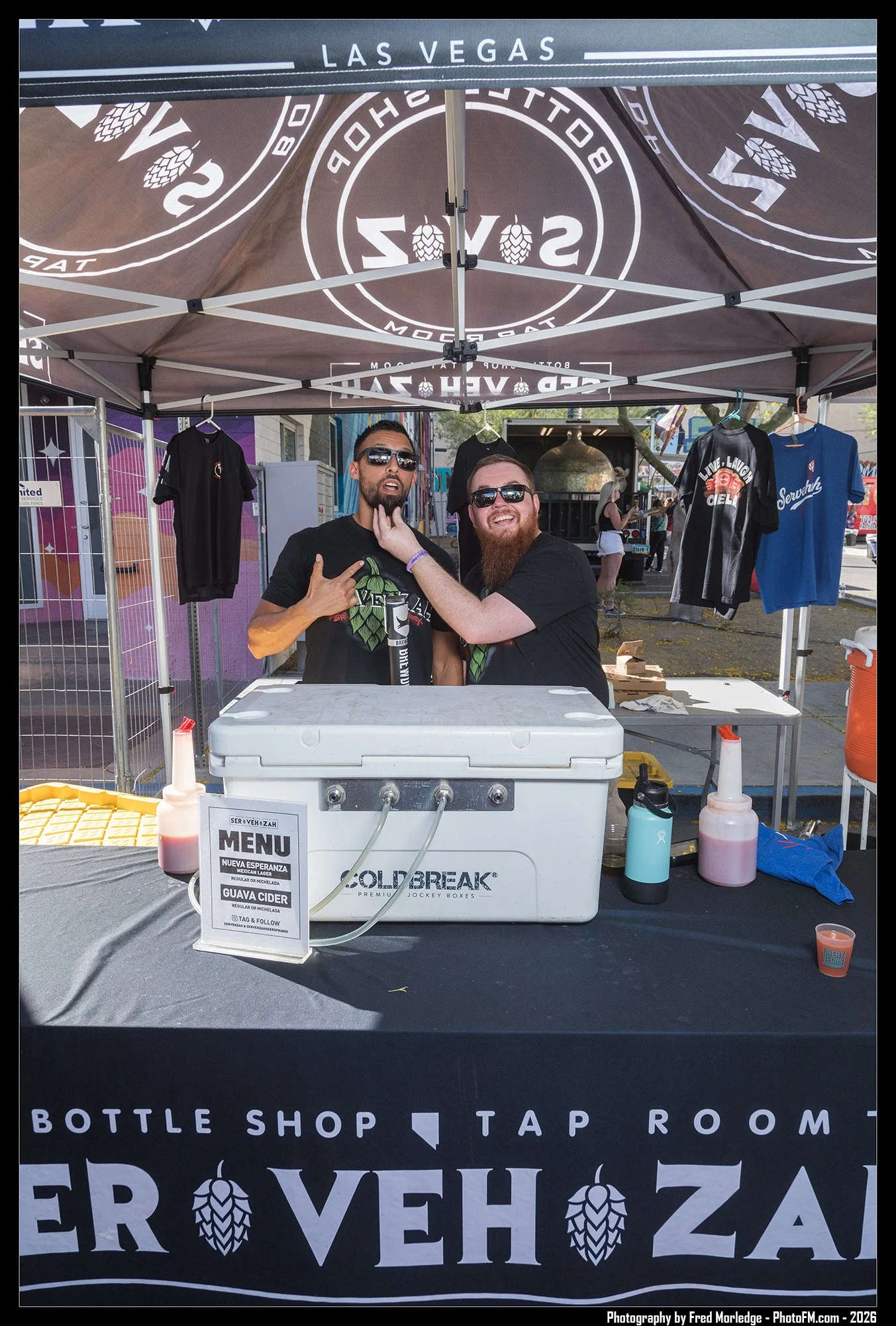 Two men with sunglasses at a bottle shop stand under a tent with the words "Las Vegas" and beer hop graphics on it. They are smiling, with one touching the other's beard, and behind them are clothing items hanging on a rack. There is a cooler with a 