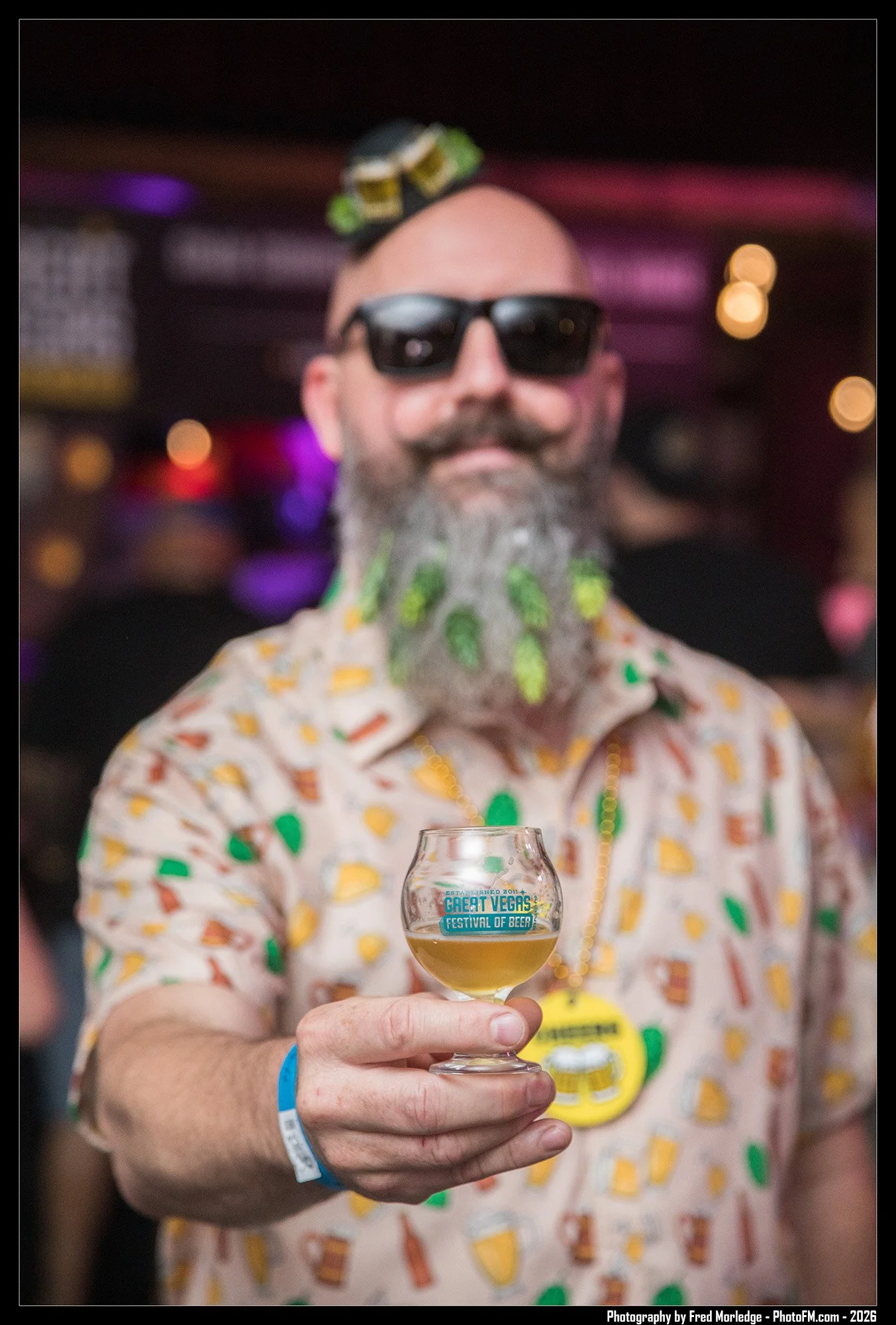 A man with a large gray beard and bald head wearing black sunglasses and a colorful shirt holding a small glass of beer at the Great Vegas Festival of Beer, with a blurred background of purple and orange lights.