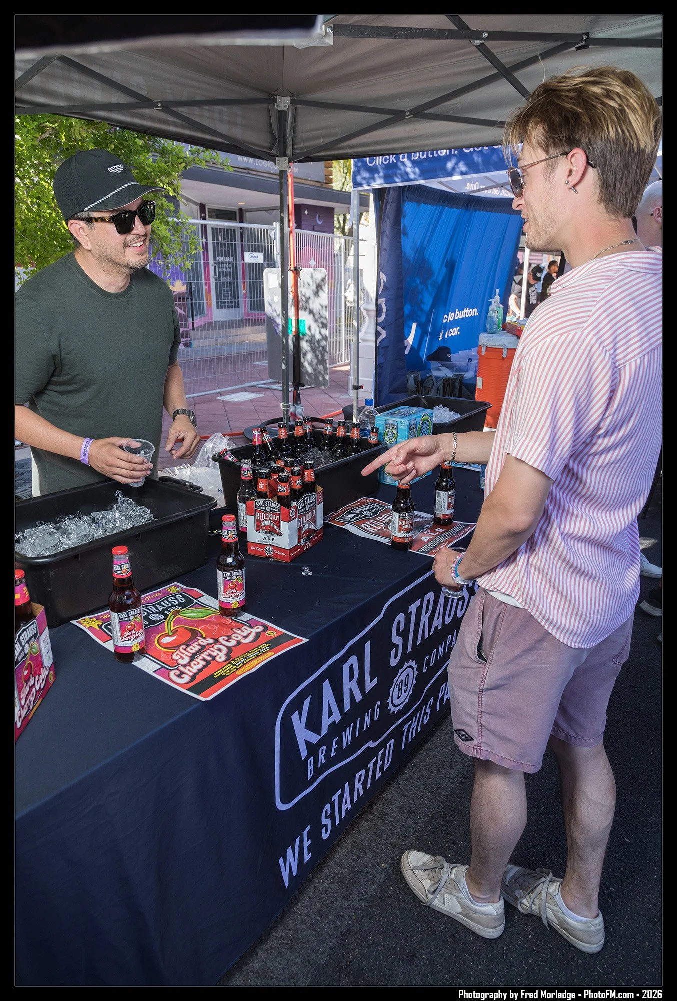 A young man wearing sunglasses, a striped pink and white shirt, and beige shorts is at a booth selling Karl Strauss Brewing Company beers at an outdoor event. He is holding a bottle of beer and engaging with a smiling man behind the booth, who is als