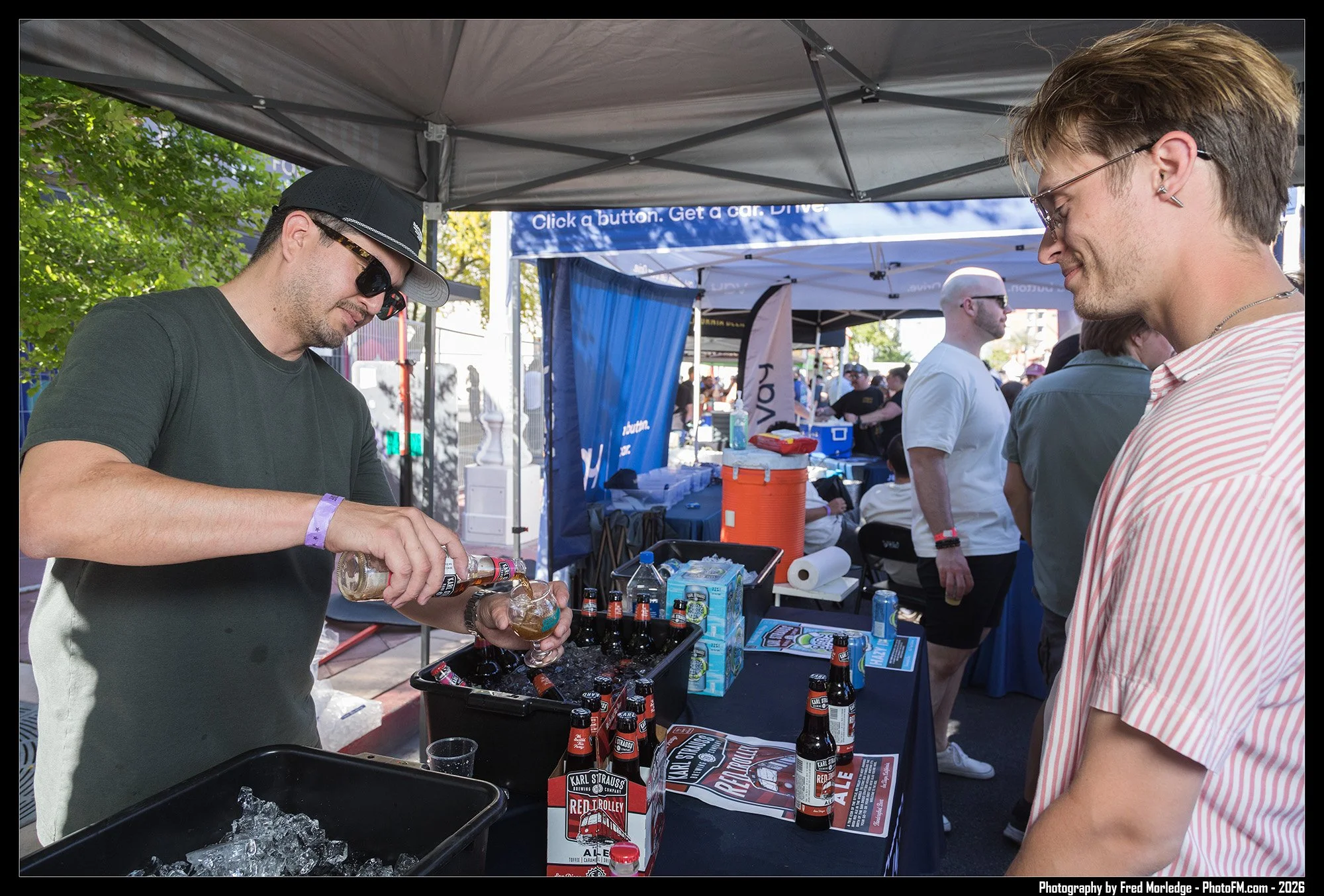A man wearing sunglasses and a black hat pouring beer into a glass at an outdoor event booth, with another man in a striped shirt smiling nearby. The booth has various beer bottles and promotional materials on the table, and there are tents and peopl