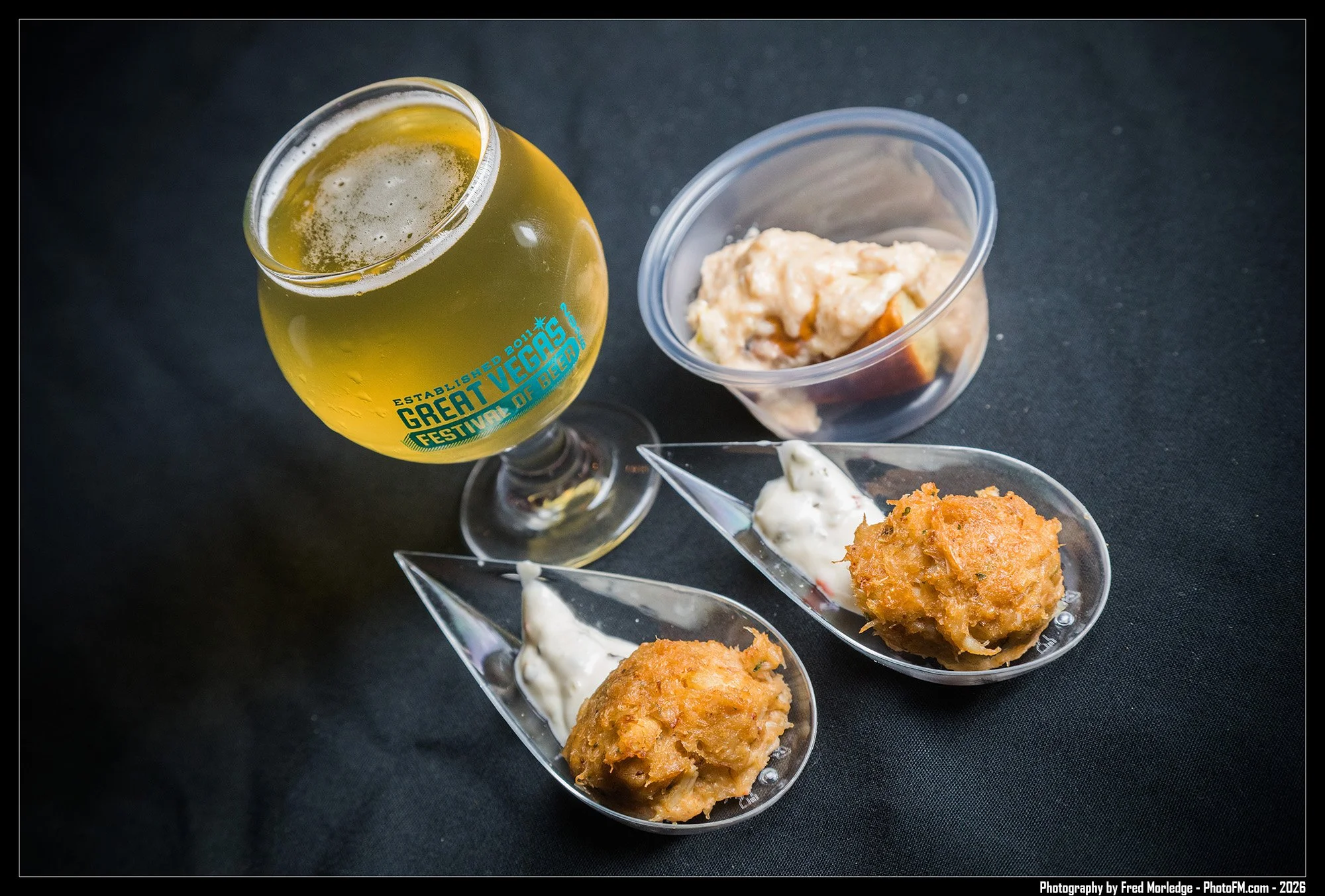 A glass of beer and several appetizers served in glass dishes on a black tablecloth. The beer glass has a logo that reads "Great Vegas Festival of Beer".