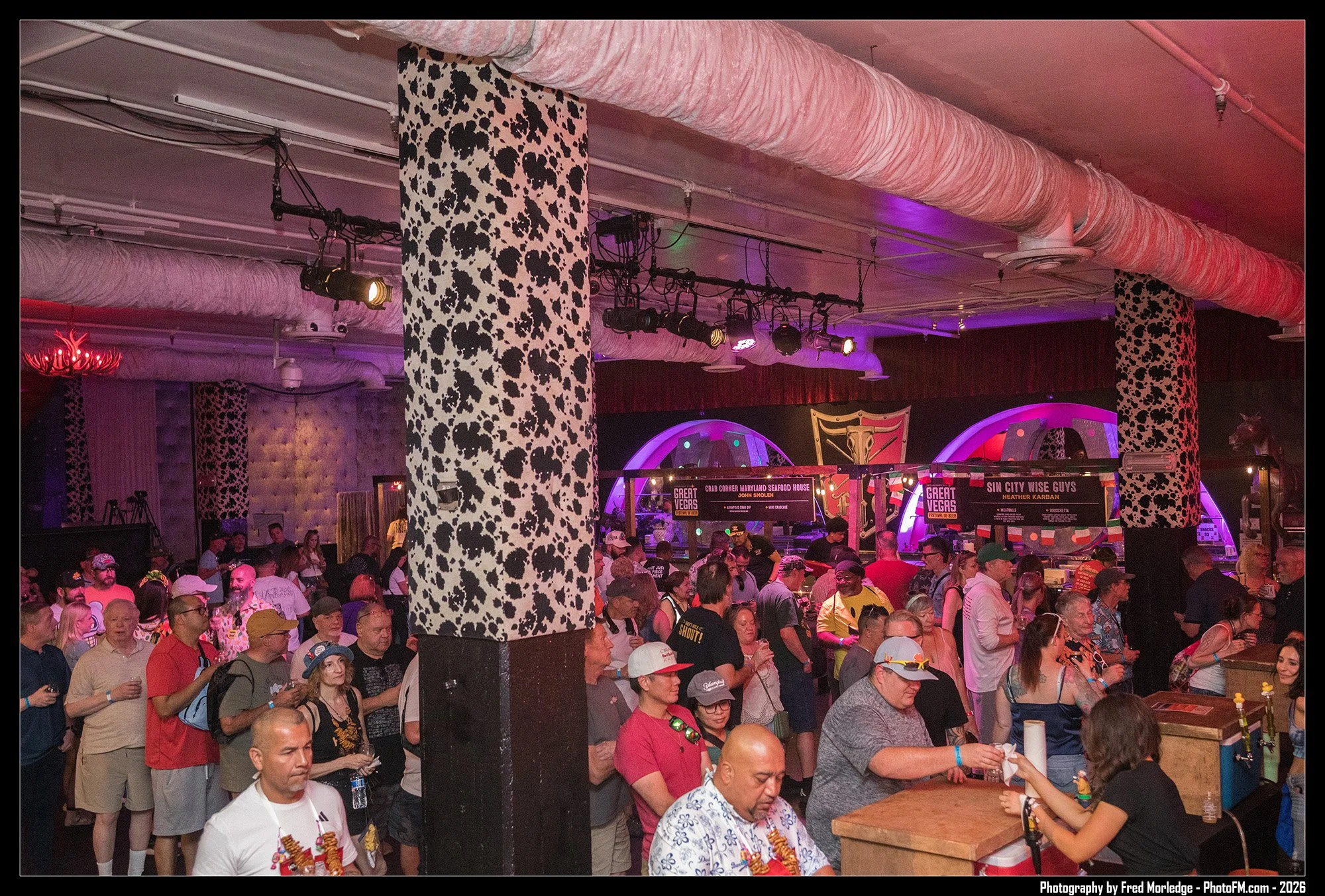 Crowd of people at a food festival with banners for crab corner Maryland seafood house and Sin City Wise Guys, with a woman wearing a gold necklace in the foreground and a large cow print column.