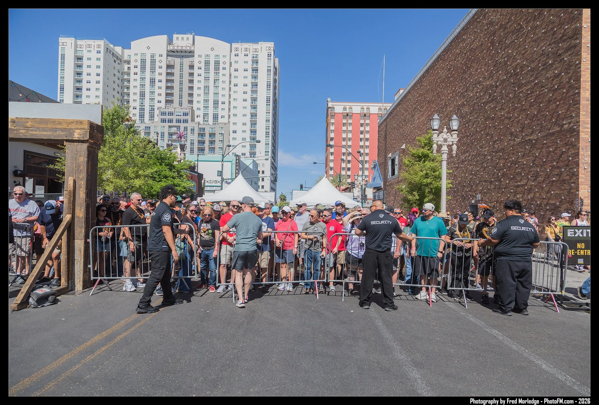 Line of people gathered behind metal barricades on city street, security personnel in black shirts standing in front, tall buildings in the background, clear blue sky.