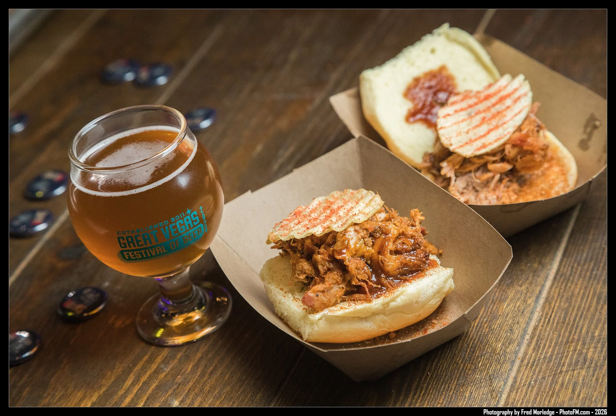 A glass of beer and two small sandwiches with pulled pork, barbecue sauce, and sandwich bread on a wooden table.