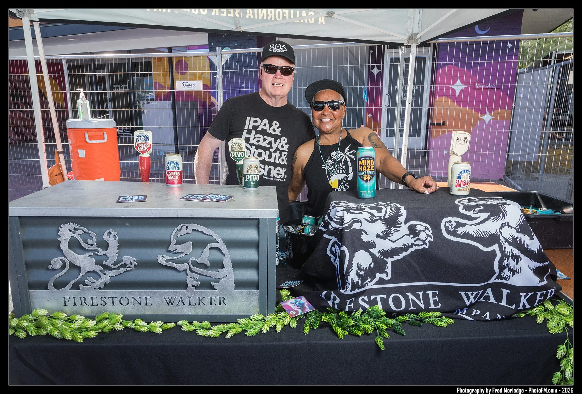 Two people standing behind a table at an outdoor event, with a metal fence and colorful mural in the background. The table displays cans of Firestone Walker beer and a black cloth with the Firestone Walker logo. Both people are wearing sunglasses, sm