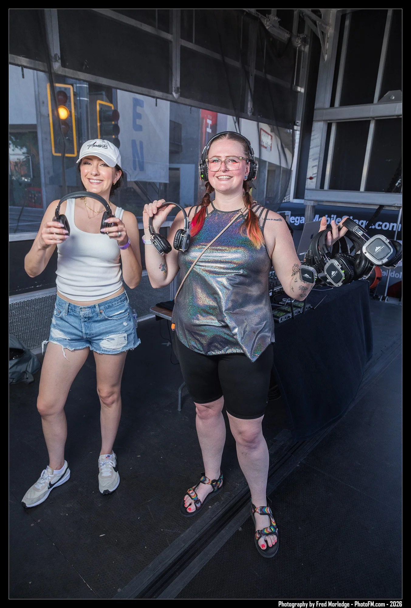 Two women standing inside a glass-walled area, holding headphones and smiling at the camera, with DJ equipment in the background.