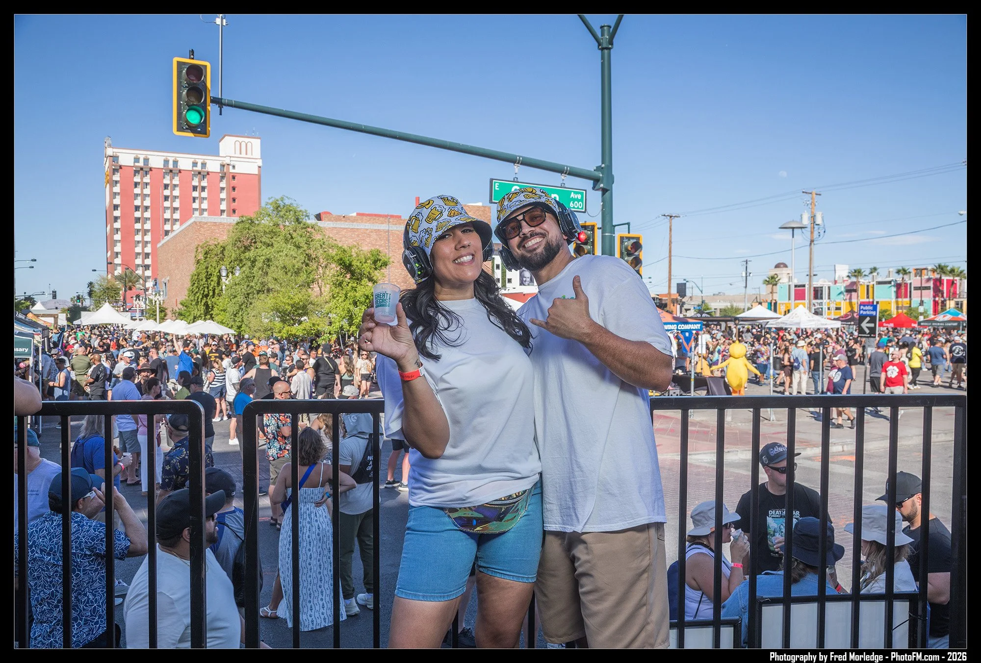 Two people standing in front of a crowd at an outdoor event, smiling and wearing matching hats and headsets; woman holding drinks. Background includes tents, trees, and buildings.