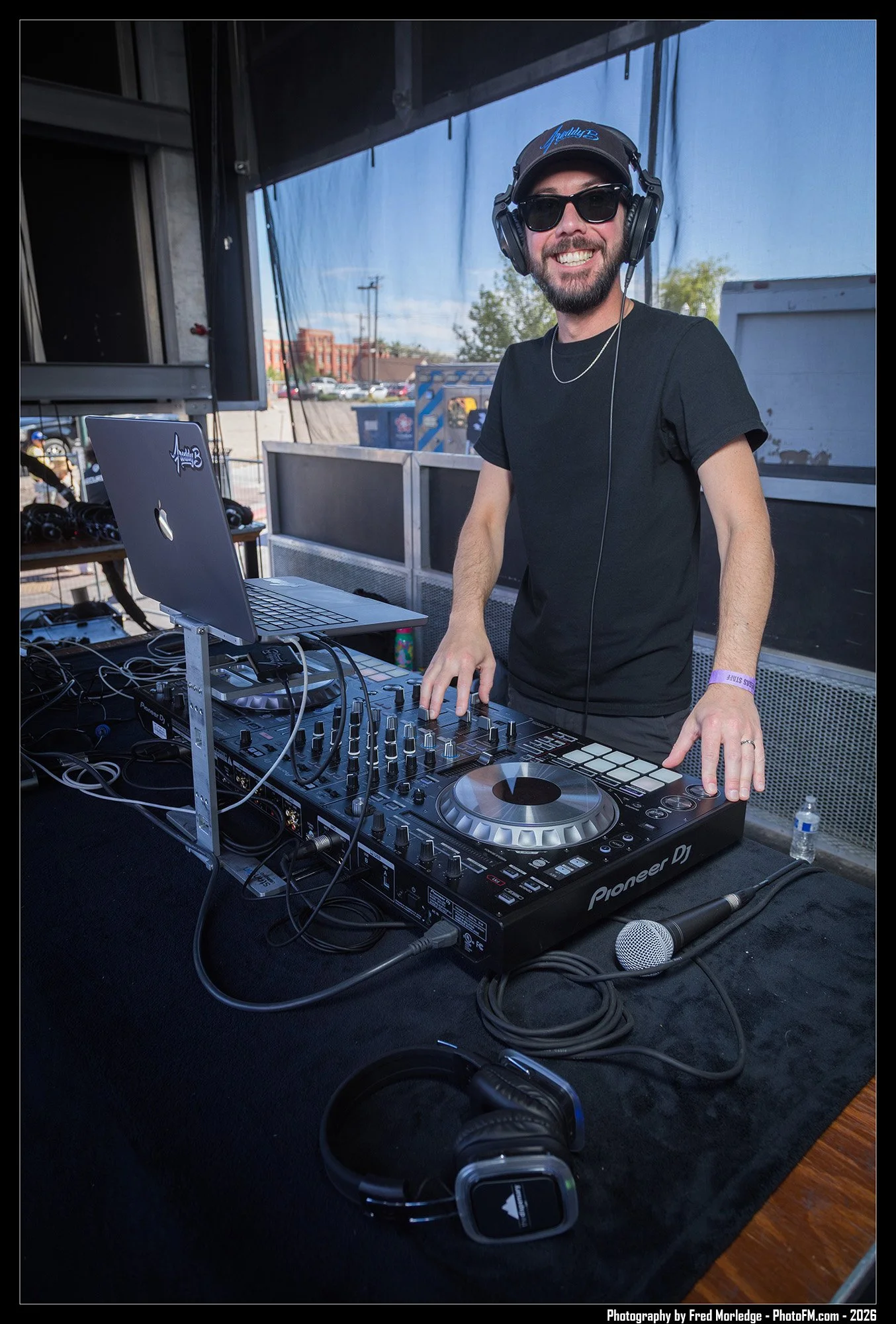 A DJ with a beard wearing sunglasses, a baseball cap, and headphones, standing behind a DJ controller and a laptop at an outdoor event, smiling at the camera.