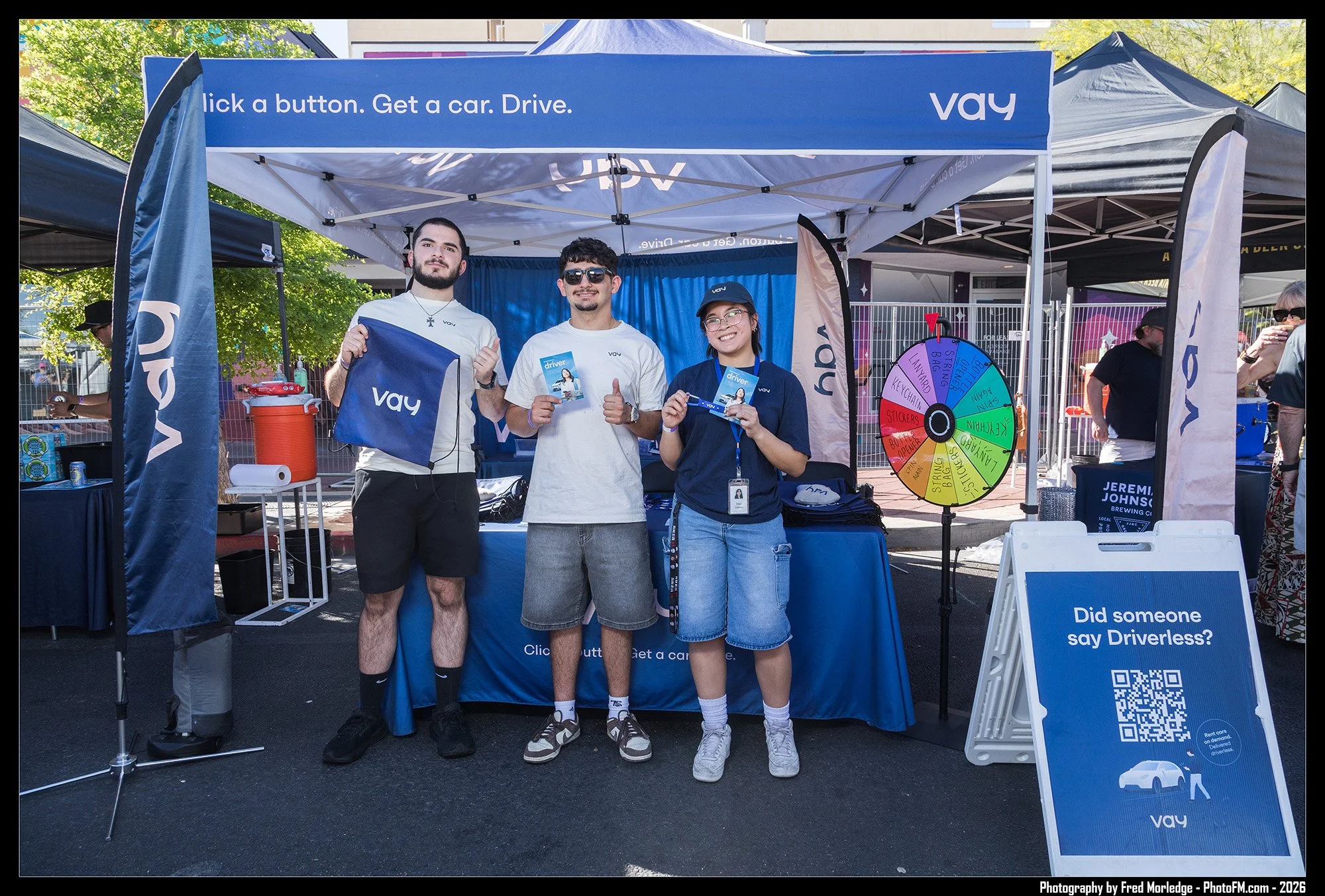 Three people standing in front of a booth promoting driverless cars at an outdoor event. They are holding promotional materials and smiling. The booth has a blue banner that reads 'click a button. Get a car. Drive.' and a spinning wheel game with var