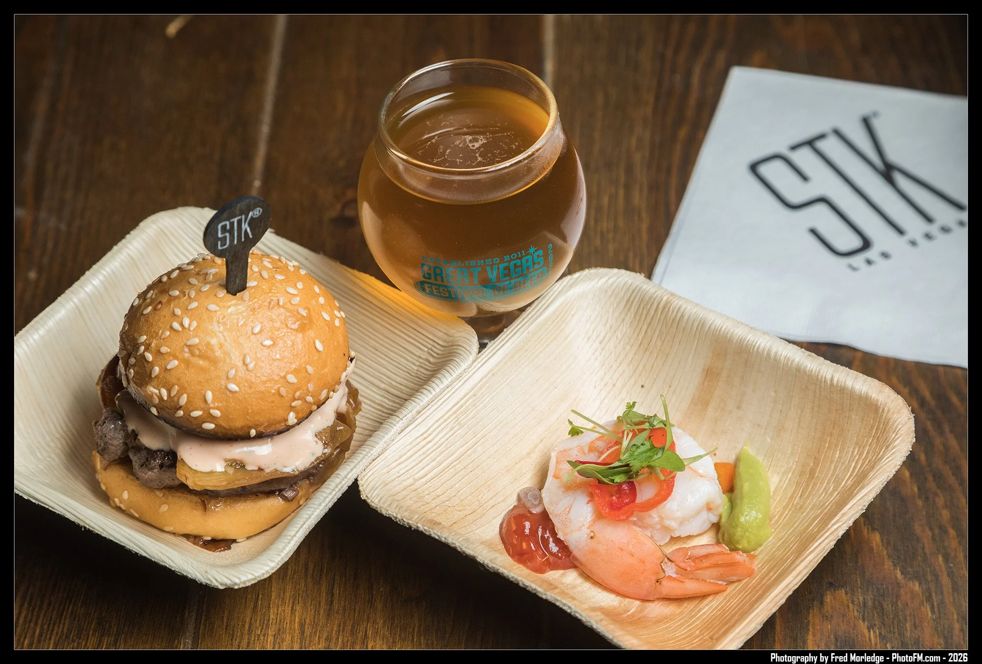 A sesame seed burger with beef patty, lettuce, and sauce on a bamboo plate, a shrimp and vegetable dish with lime and cilantro, and a glass of beer with a "Great Vegas Festival of Beer" logo on a wooden table.