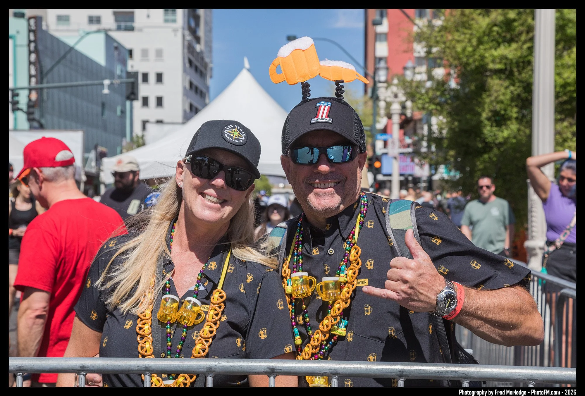 Two smiling people at an outdoor event, wearing black shirts with gold beer mug patterns, multicolored beads, and rainbow glasses. The man has a beer mug hat on his head and a big smile, giving a thumbs up. The woman is wearing sunglasses and a black