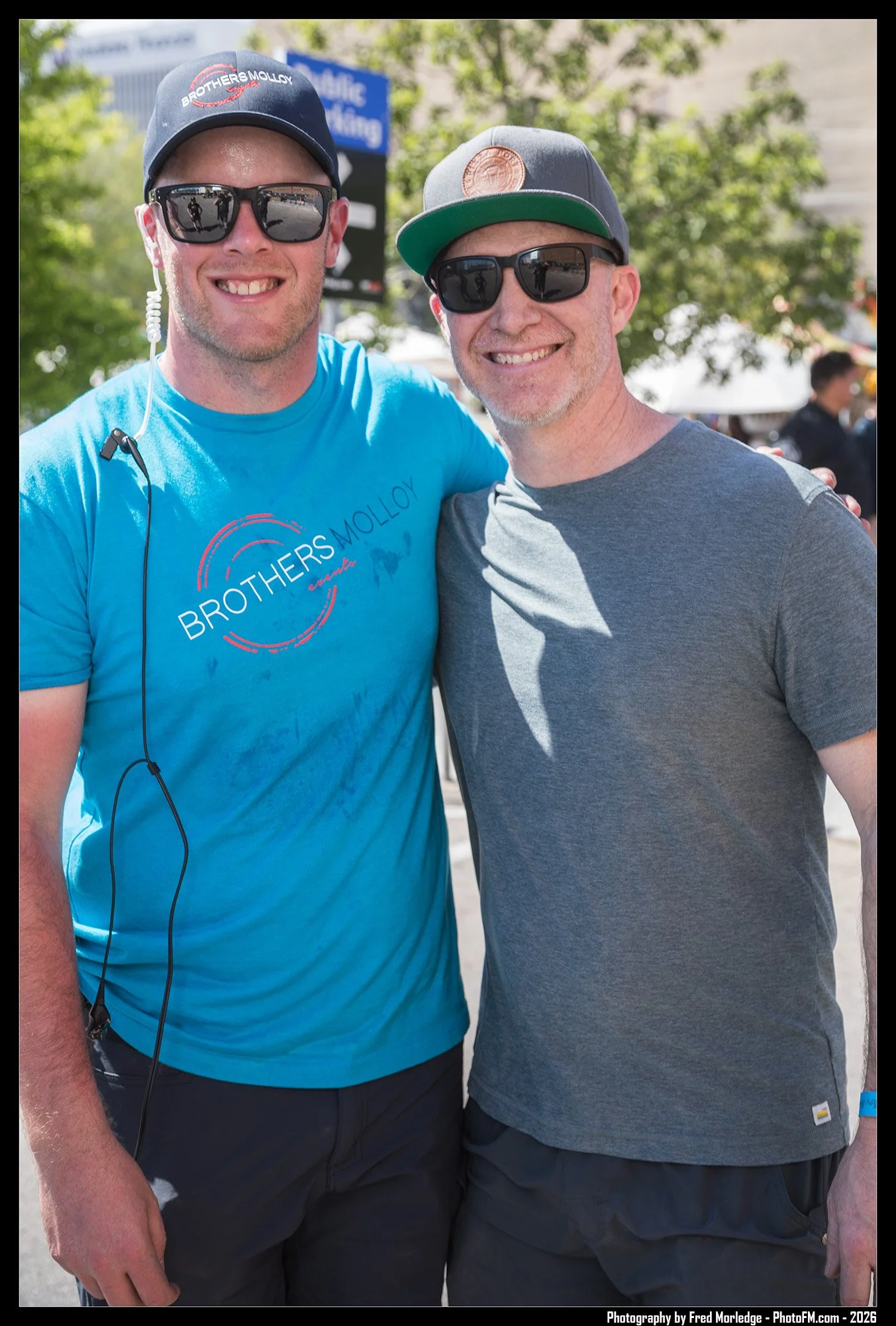 Two men standing close together outdoors, smiling at the camera. They are wearing sunglasses, baseball caps, and casual t-shirts. One man's shirt has the text 'Brothers Molloy' on it. The background has green trees and a few people.