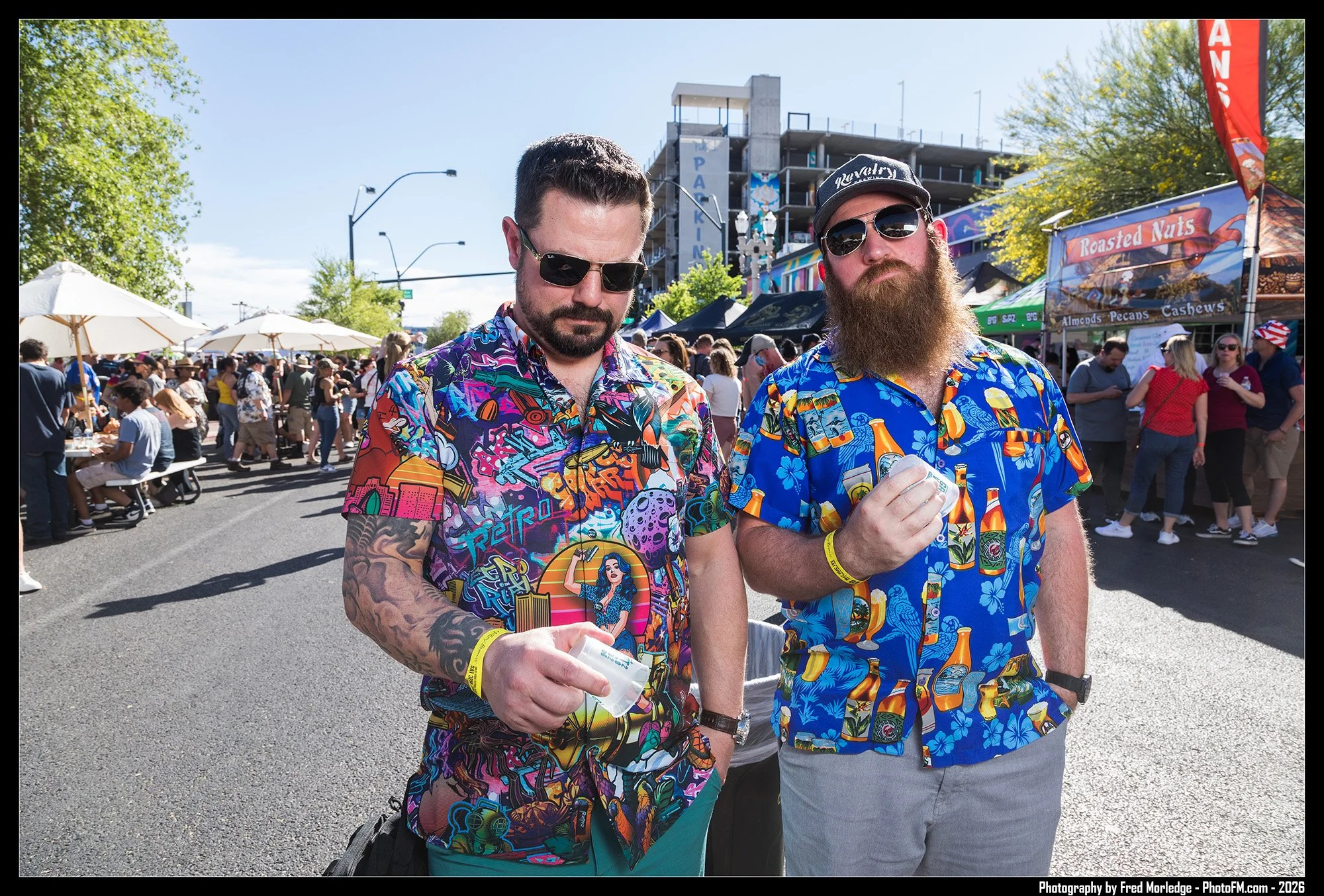 Two men wearing colorful Hawaiian shirts and sunglasses at an outdoor event or fair. One man has a full beard and cap, and the other with tattoos on his arm is holding sunglasses in his hand. The background shows booths, tents, and a crowd.