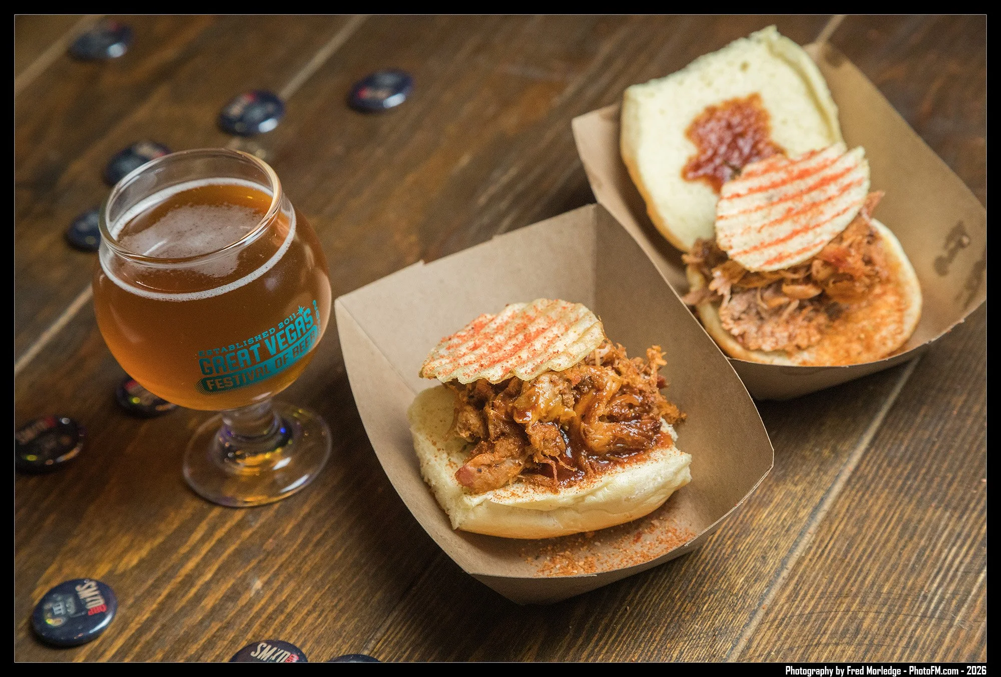 A glass of amber-colored beer with a blue label, two pulled pork buns with spicy sauce and slice of bread, and a cardboard box containing two pulled pork sandwiches with buns, shredded pork, sauce, and a slice of bread, on a wooden table.