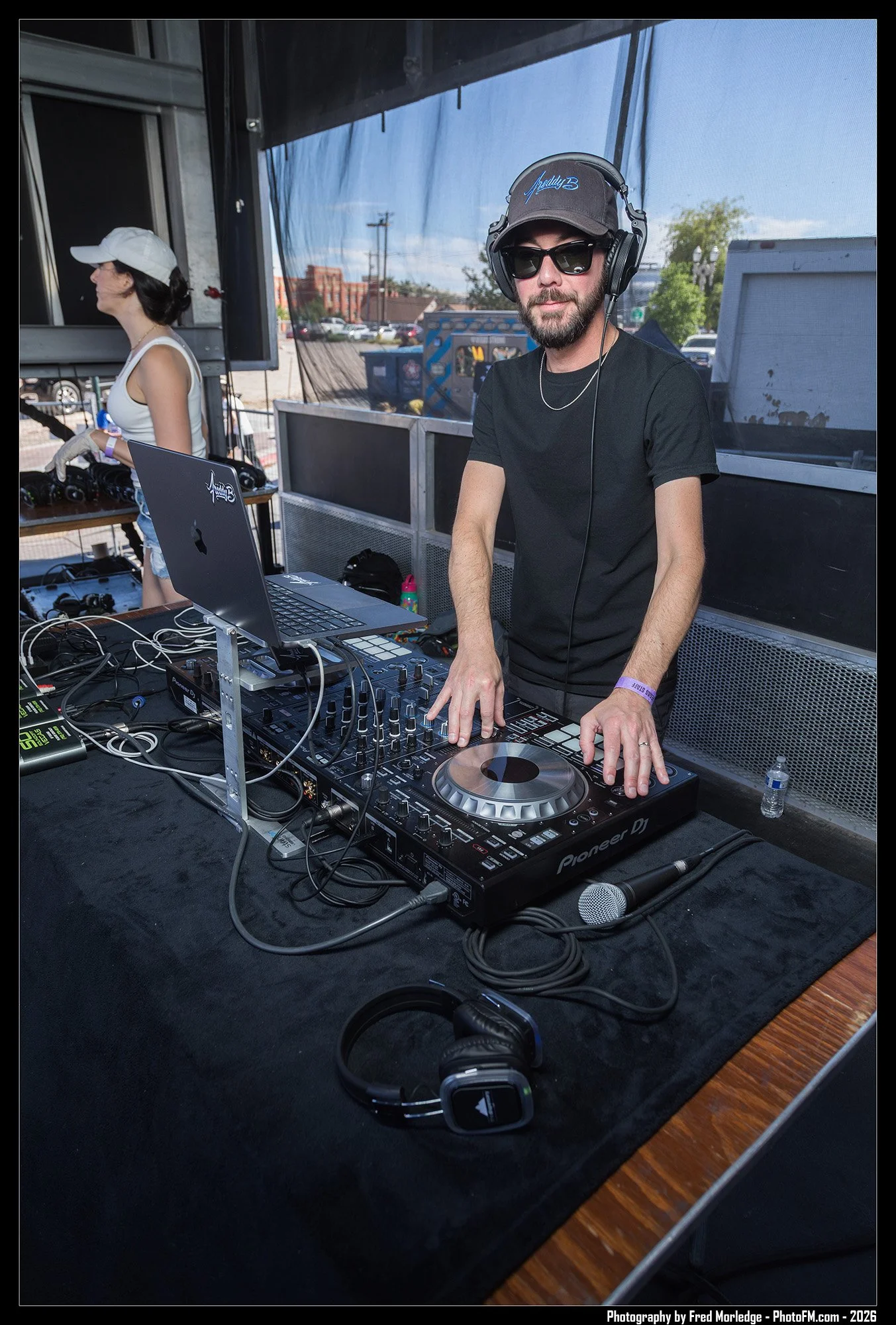 A DJ man with a beard, wearing sunglasses, a baseball cap, and a black t-shirt, is DJing at an outdoor event. He is standing behind a DJ deck with a laptop and other equipment. A woman in a white tank top and white cap is in the background, standing 