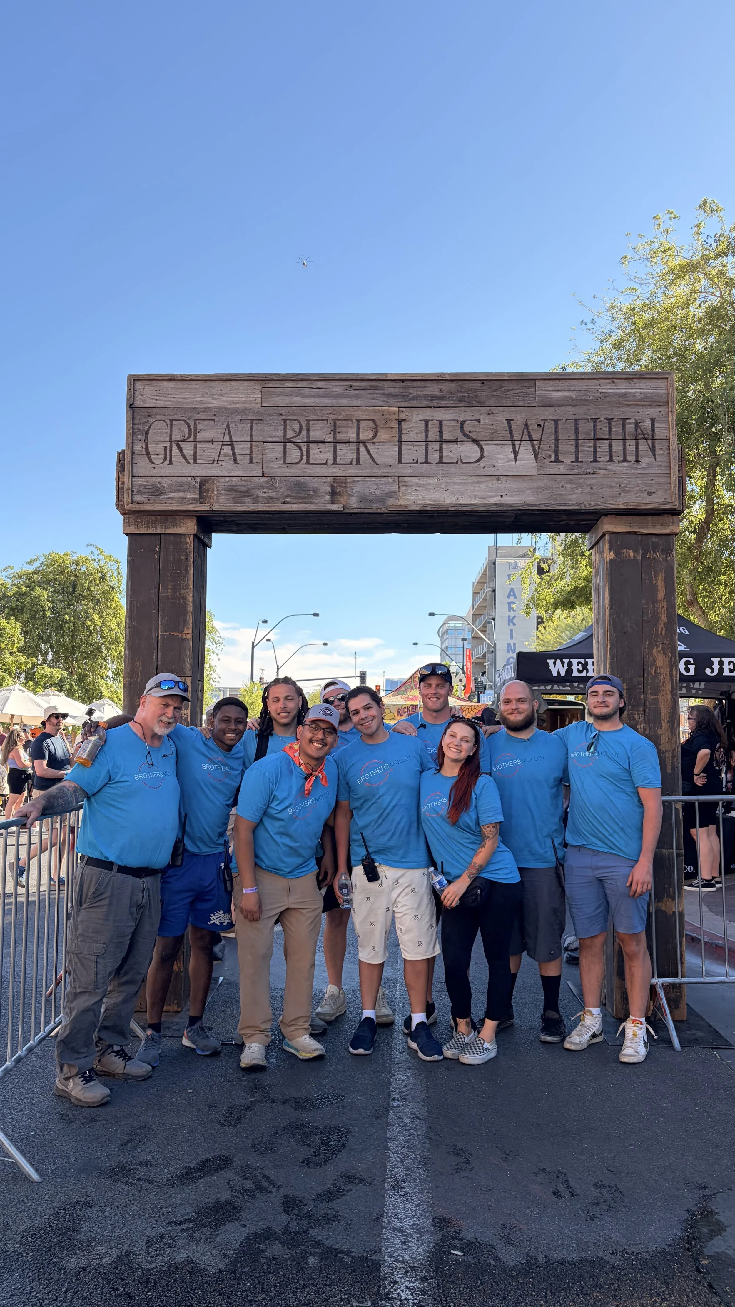 Group of people smiling and posing for a photo beneath a wooden arch with the phrase "Great Beer Lies Within" engraved on it, at an outdoor event on a sunny day.