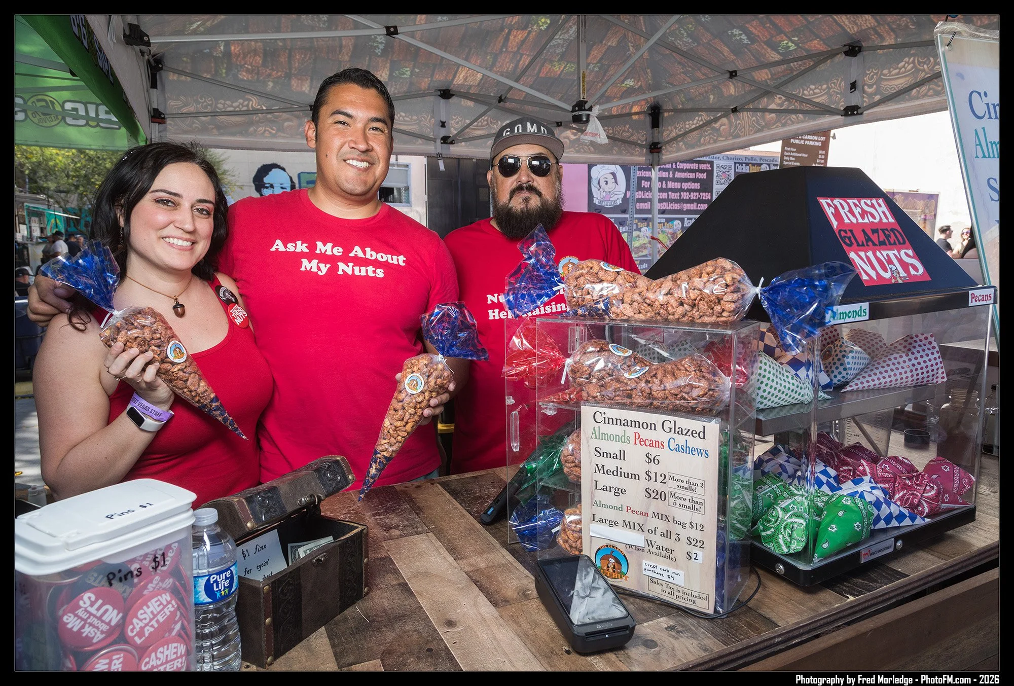 Three people standing behind a nut stand at an outdoor event. The woman on the left is holding a bag of candied nuts and smiling. The man in the middle is wearing a red shirt that says 'Ask Me About My Nuts' and is smiling. The man on the right has a
