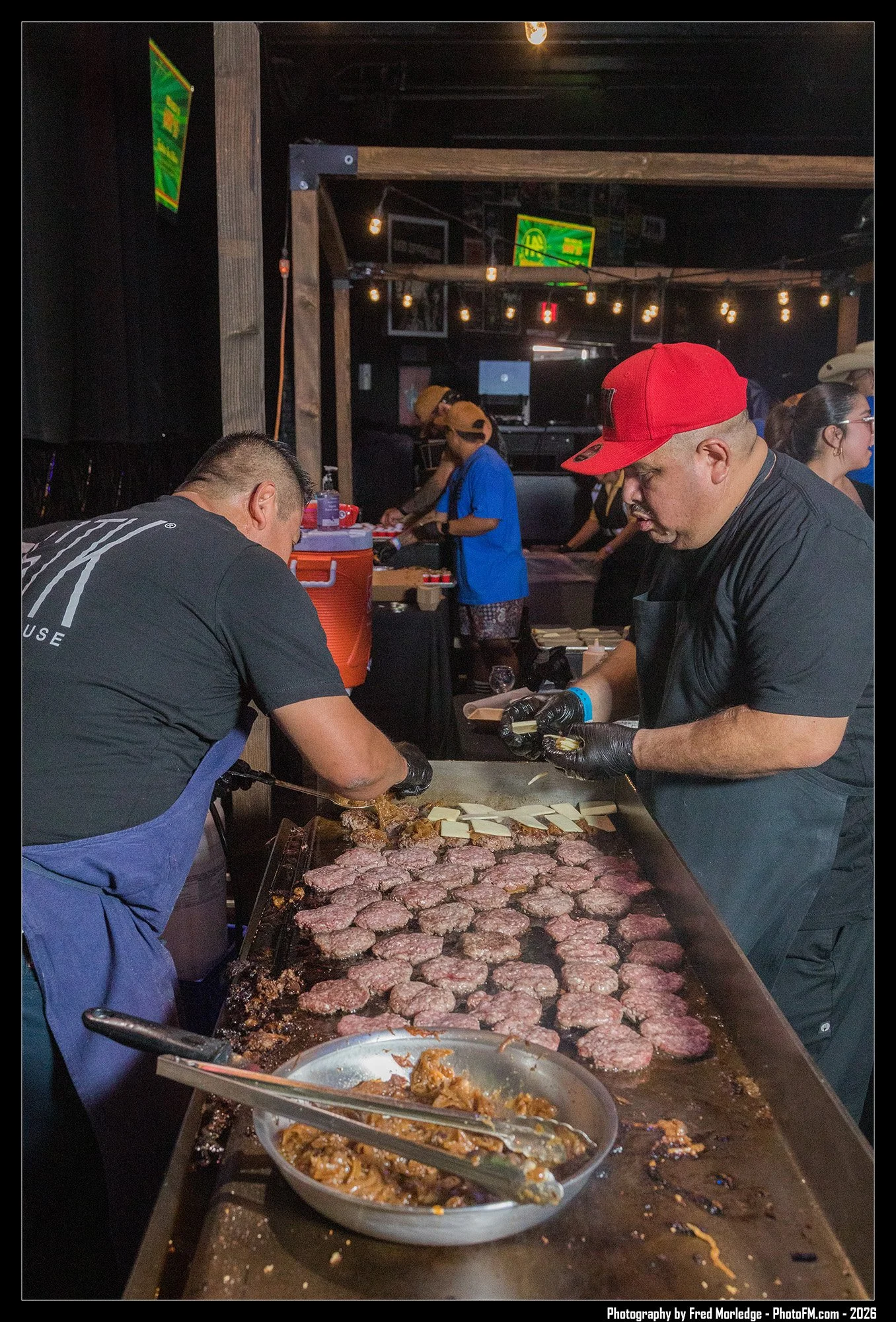 Two men are cooking hamburger patties on a large flat-top grill, with one of them flipping the patties and the other preparing toppings. Several other people are in the background, working at a food stand, with neon signs and string lights overhead, 