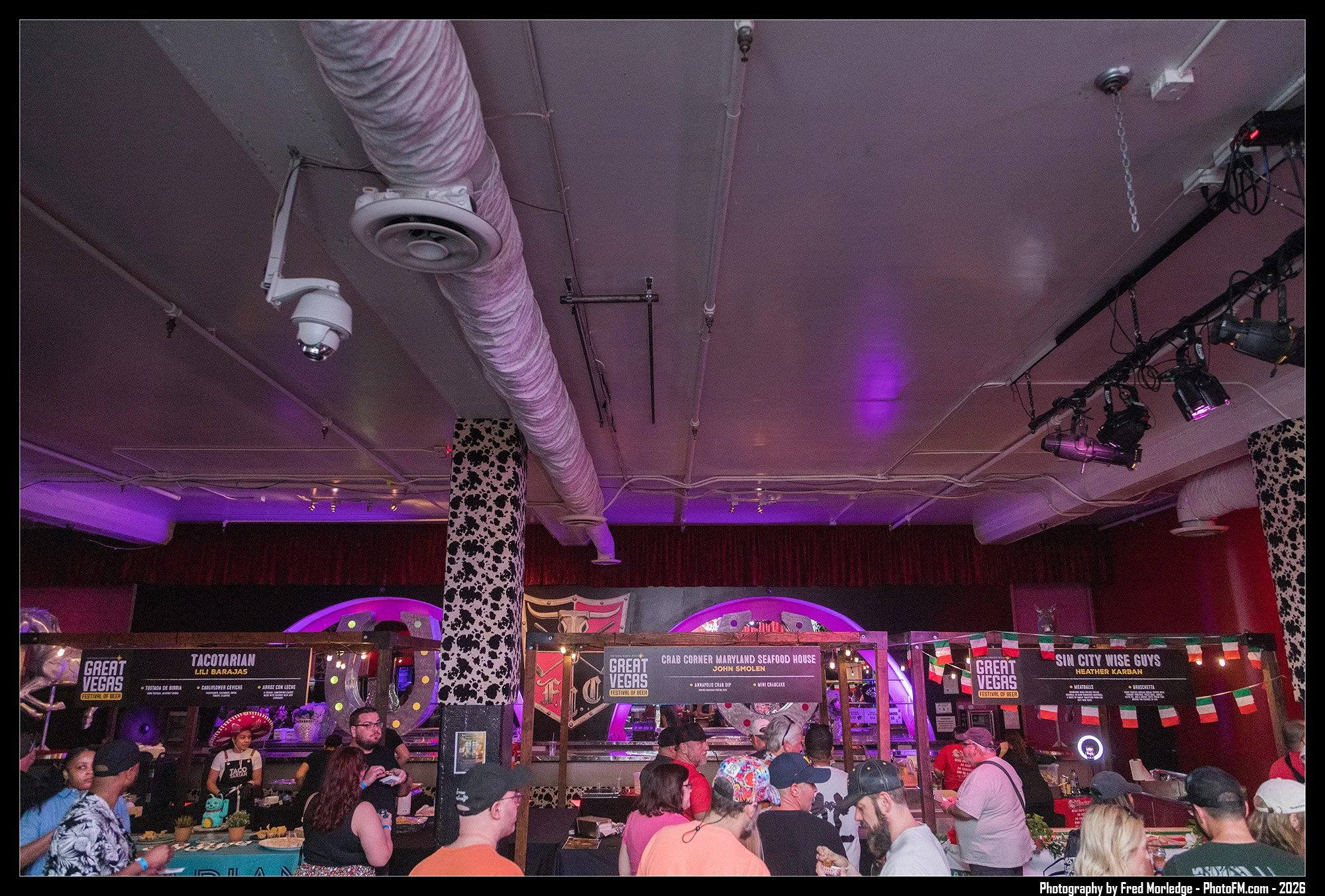 People gathered at food booths at the Great Vegas Festival of Beer, with signs displaying different food vendors and a variety of people in line and talking, inside a room with purple lighting and visible equipment on the ceiling.