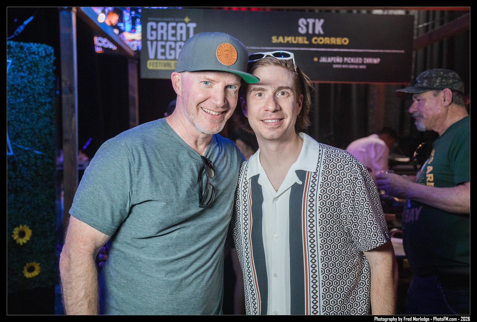 Two men are smiling and standing close together at a festivity or event. The man on the left is wearing a gray t-shirt and a gray baseball cap with an orange logo, while the man on the right is dressed in a patterned shirt with a white collar. The ba