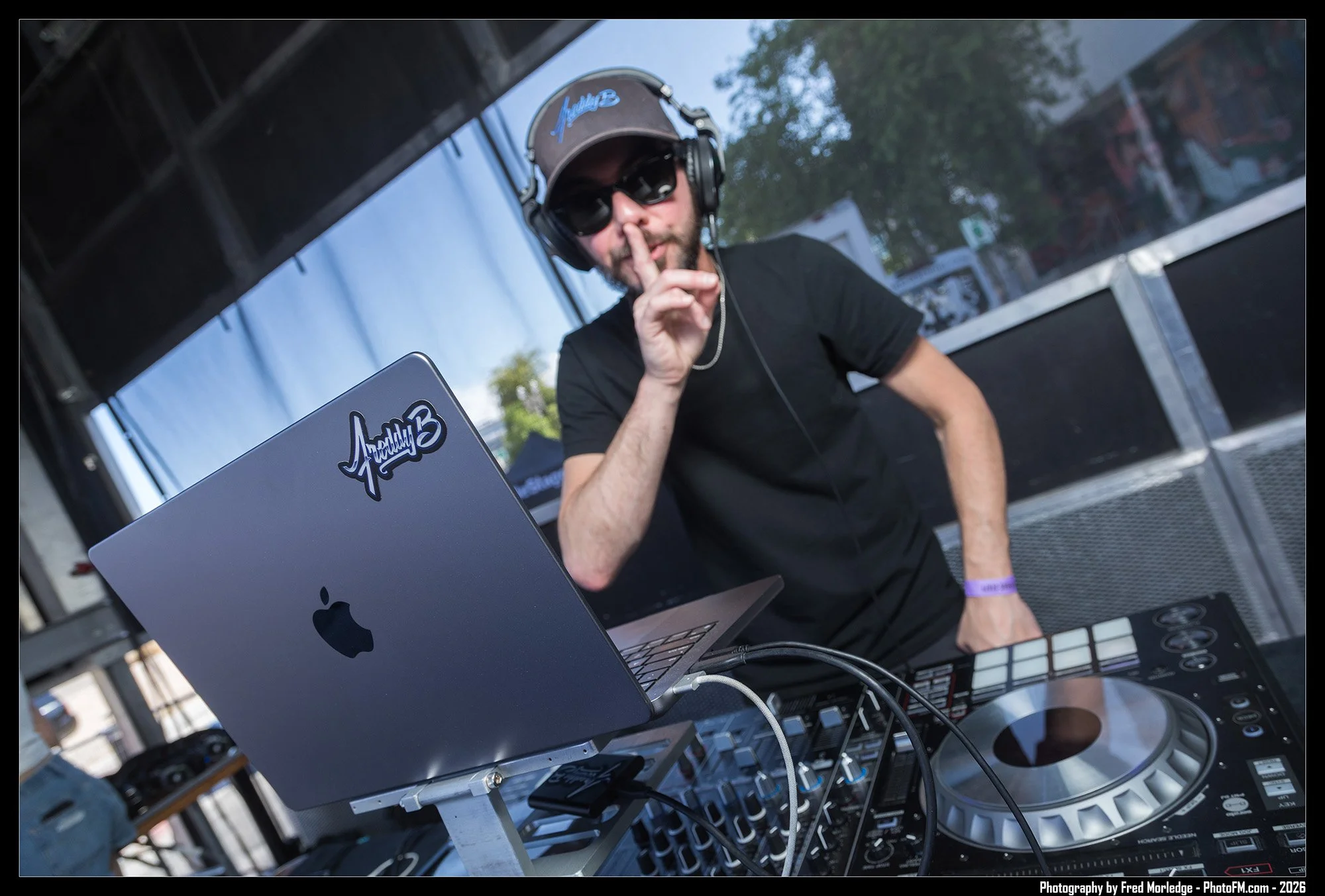 A DJ wearing sunglasses, a brown cap, and headphones, gesturing with a finger over his lips while standing behind DJ equipment and a laptop that has a sticker reading 'Pretty 3'.