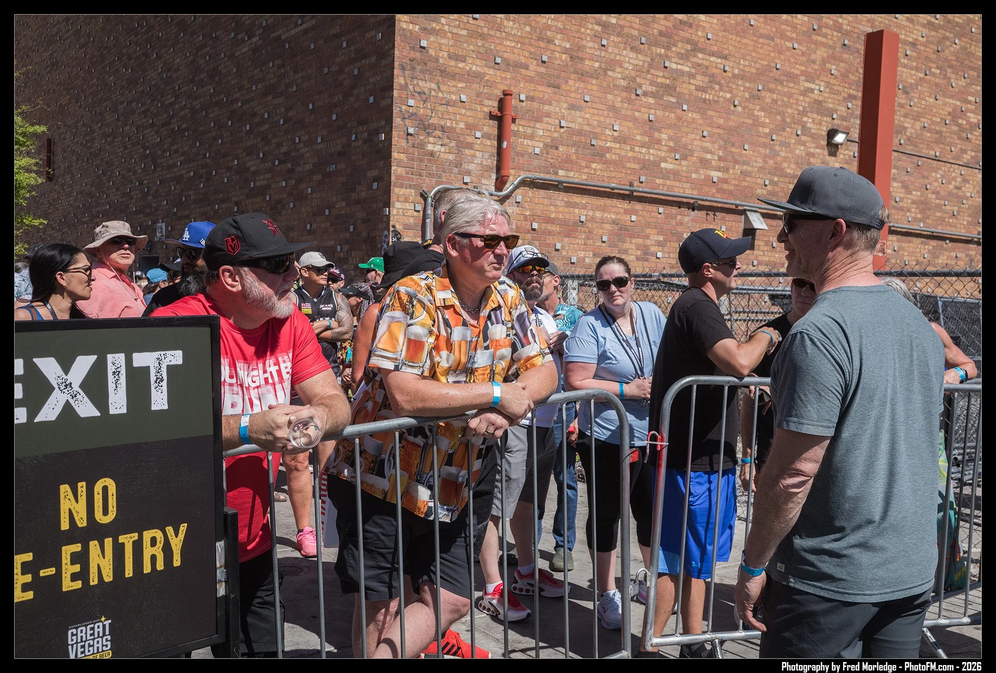 Group of people standing behind a metal barricade outside a brick building, engaging in conversation at an event or gathering.