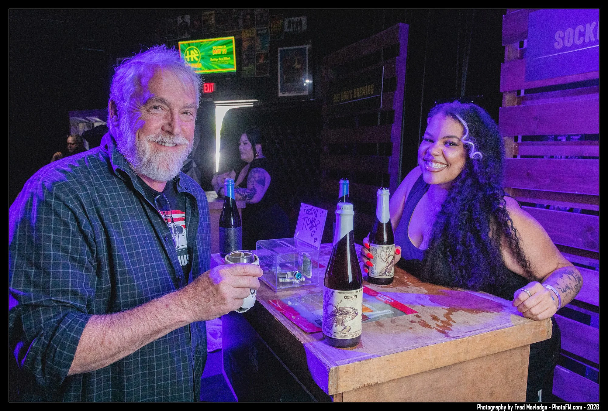 A man with white hair and a beard, wearing a plaid shirt, sitting at a bar with two bottles of beer, smiling. A woman with long curly hair and tattoos, smiling, sitting across from him with a beer. The background is dimly lit with purple and blue lig