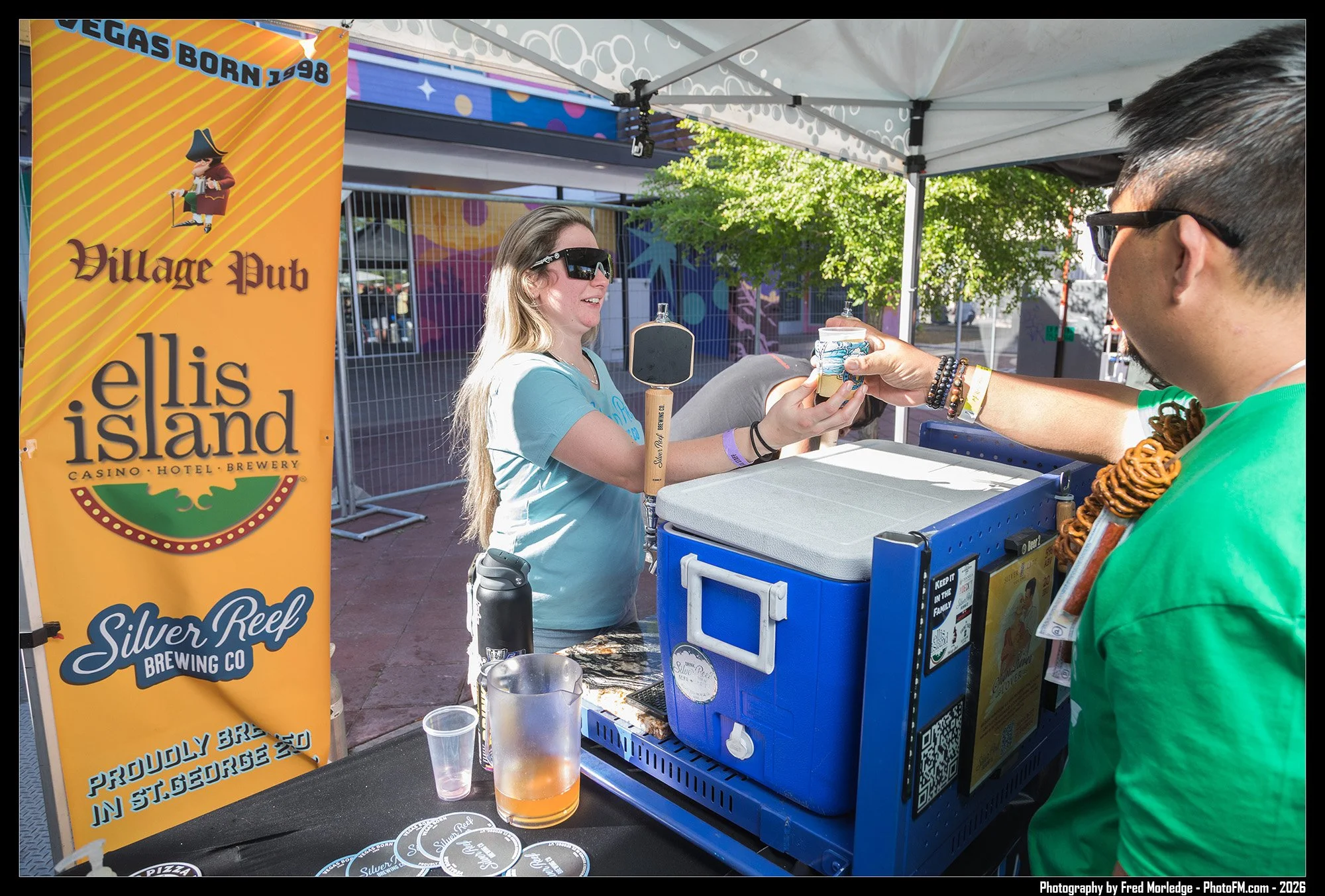 A woman serving beer at a booth with a sign reading 'Villlage Pub', 'Ellis Island', and 'Silver Reef Brewing Co.'. She is wearing sunglasses and a blue shirt, handing a cup of beer to a person in a green shirt. The booth is set up outdoors under a te