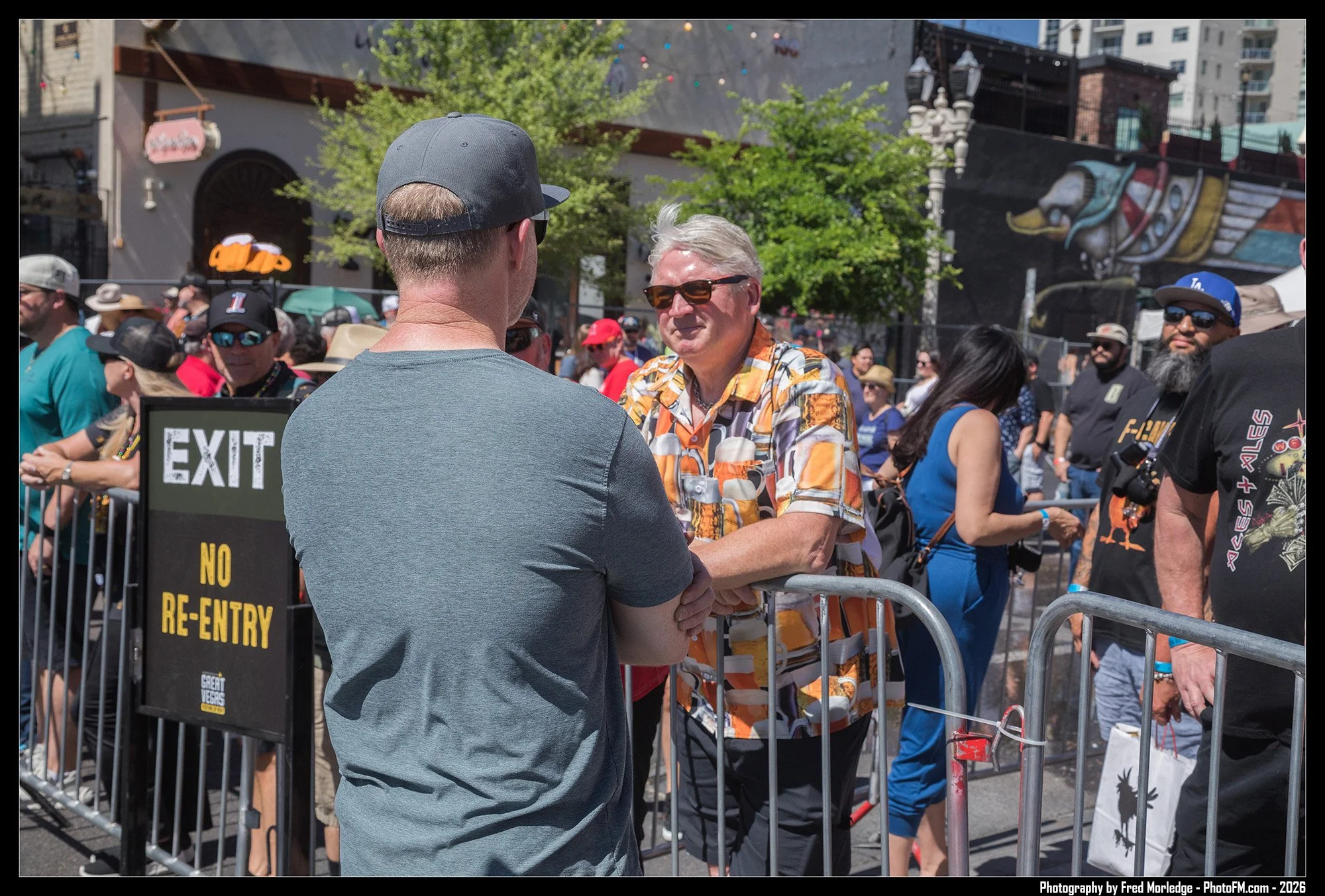People gathering behind metal barricades at an outdoor event, with a man wearing a gray cap and sunglasses engaged in conversation with another man in a colorful Hawaiian shirt. There are various individuals wearing hats and sunglasses, and a mural o