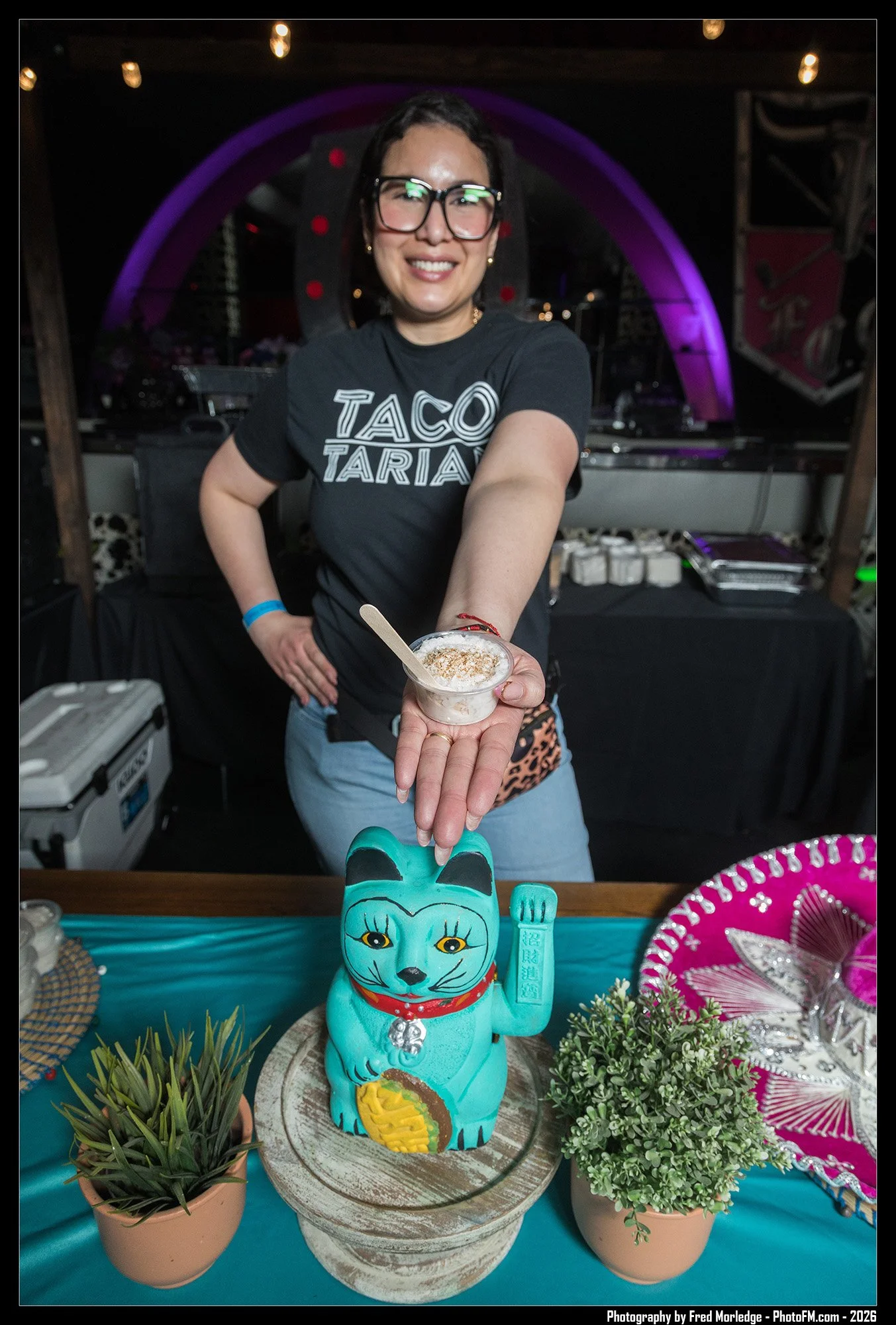 A smiling woman wearing glasses and a black T-shirt with the words "Taco Taria" is holding a small dessert cup. In front of her, a table display features a turquoise ceramic cat figurine and various plants and decorations, including a pink fan and sm