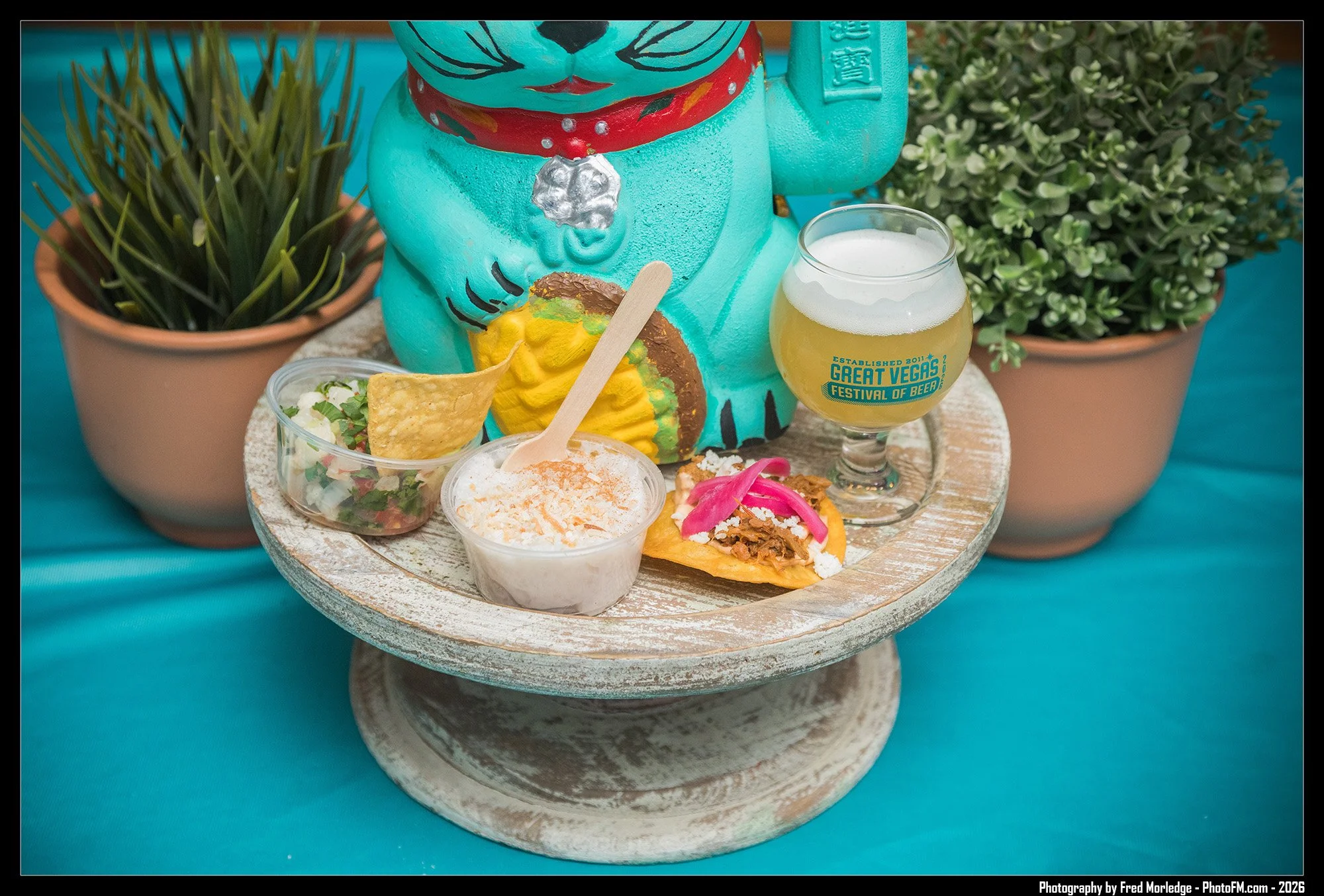 A decorative display of food and drink on a round, rustic, white wooden stand. Includes small containers of salads and toppings, a taco with pink pickled onions, a glass of beer labeled 'Great Vegas Festival of Beer,' a turquoise cat statue with a re