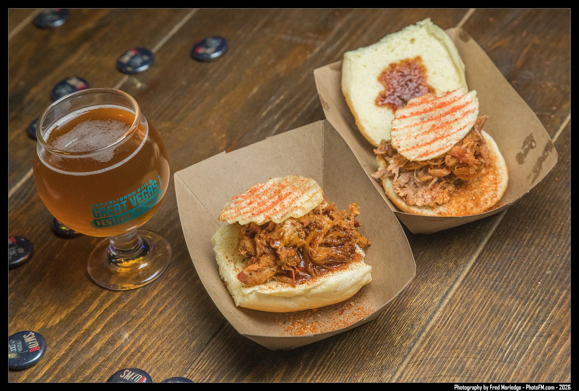 A glass of beer and two opened sandwiches with shredded meat and potato chips on top, served in paper trays on a wooden table.