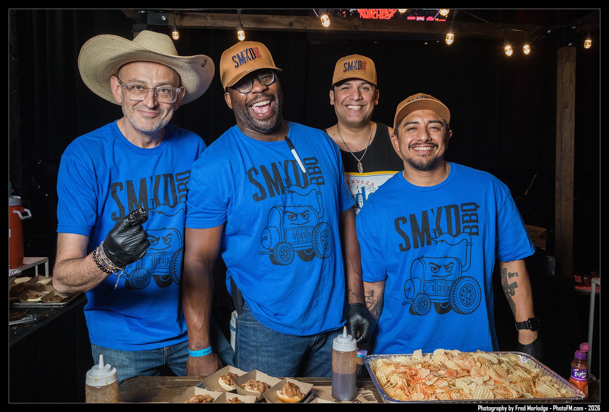Four men, wearing blue shirts with a trading robot logo, standing behind a table with food and condiments at a barbecue event.