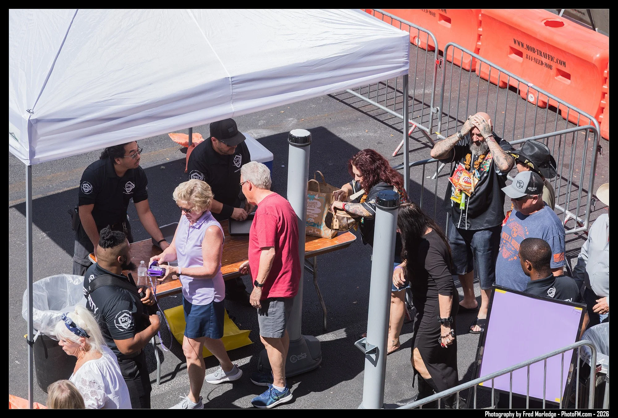 Line of people waiting at a registration or check-in booth under a white canopy tent at an outdoor event, with security personnel managing the queue and orange traffic barriers surrounding the area.