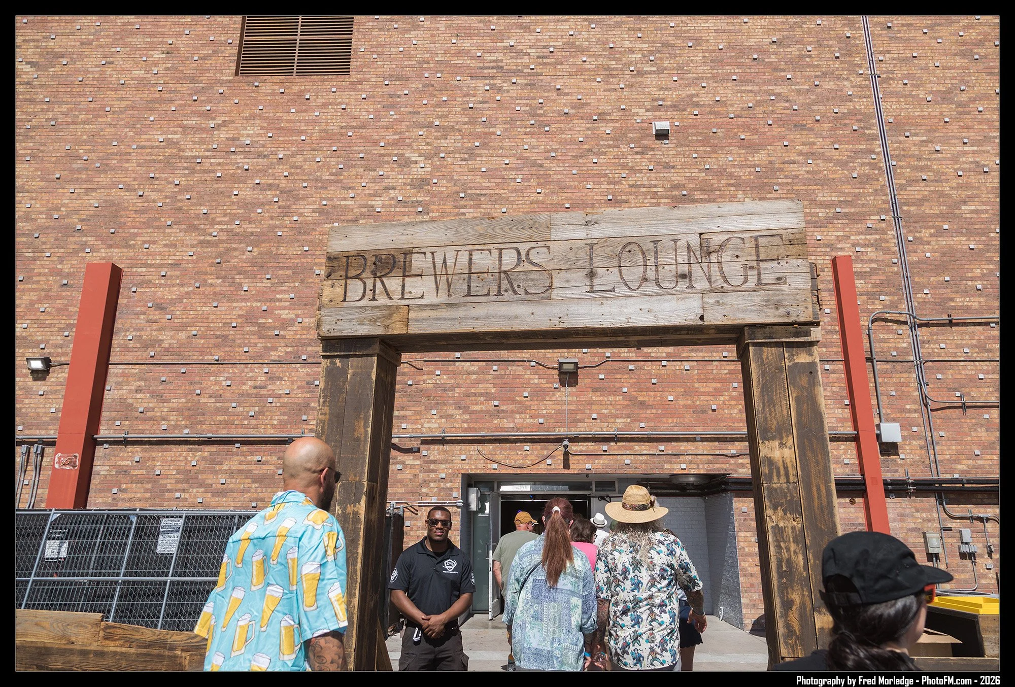 People entering the Brewers Lounge through a rustic wooden archway against a brick wall, with some wearing colorful, casual clothing and hats.