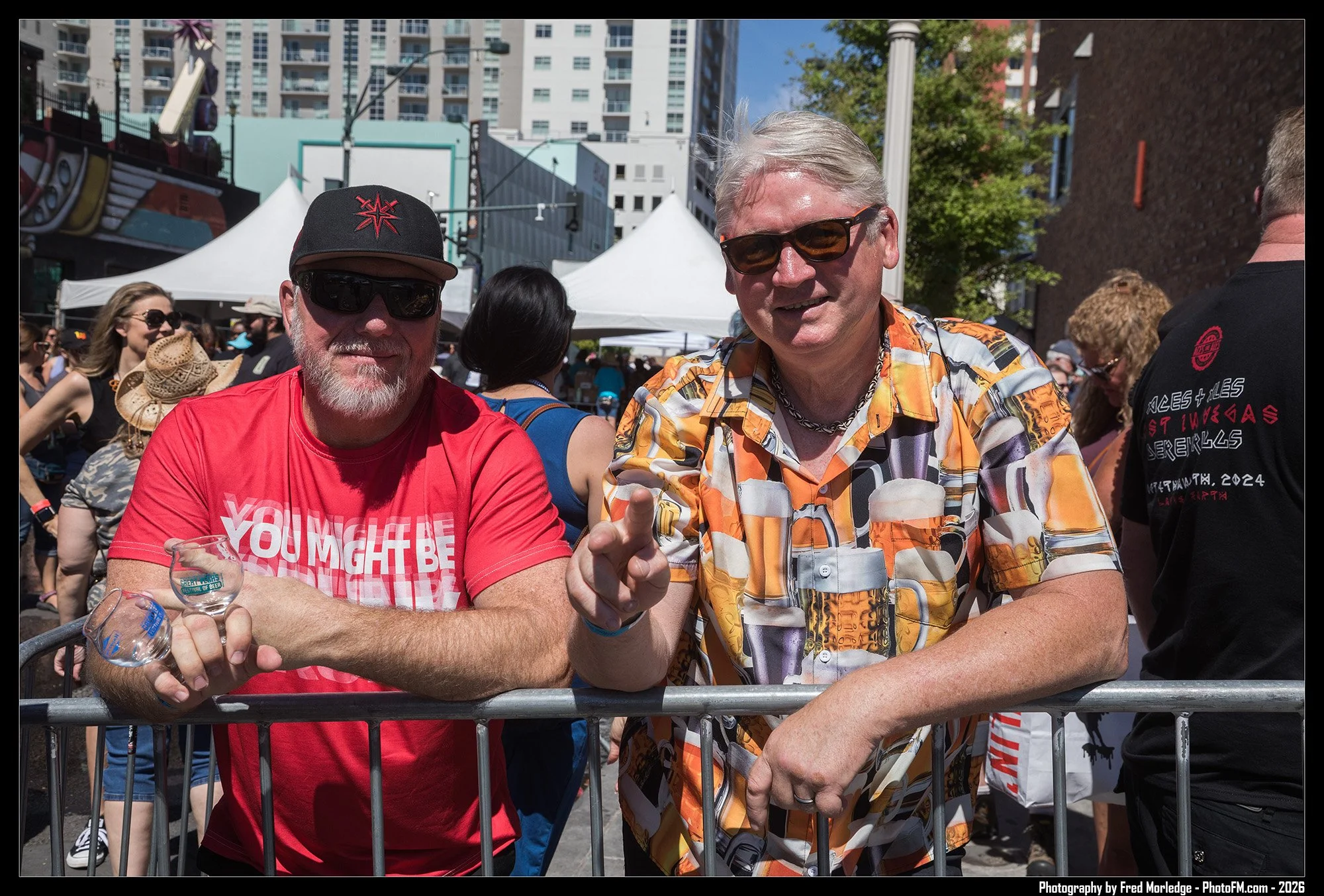 Two men with gray hair and sunglasses standing behind a metal barrier at an outdoor event, smiling and interacting. The man on the left wears a red t-shirt and a black cap, the man on the right wears a colorful patterned shirt. There are tents and a 