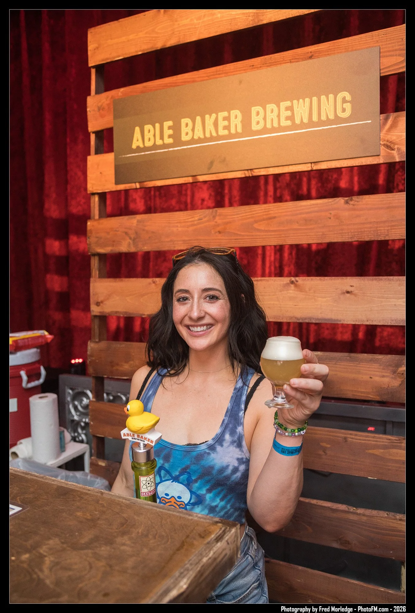 A woman with dark hair, wearing a blue tie-dye tank top and various bracelets, is smiling and holding a glass of beer with a foam head at a bar or brewery. The bar features a wooden backdrop and a sign that reads 'Able Baker Brewing' behind her, alon