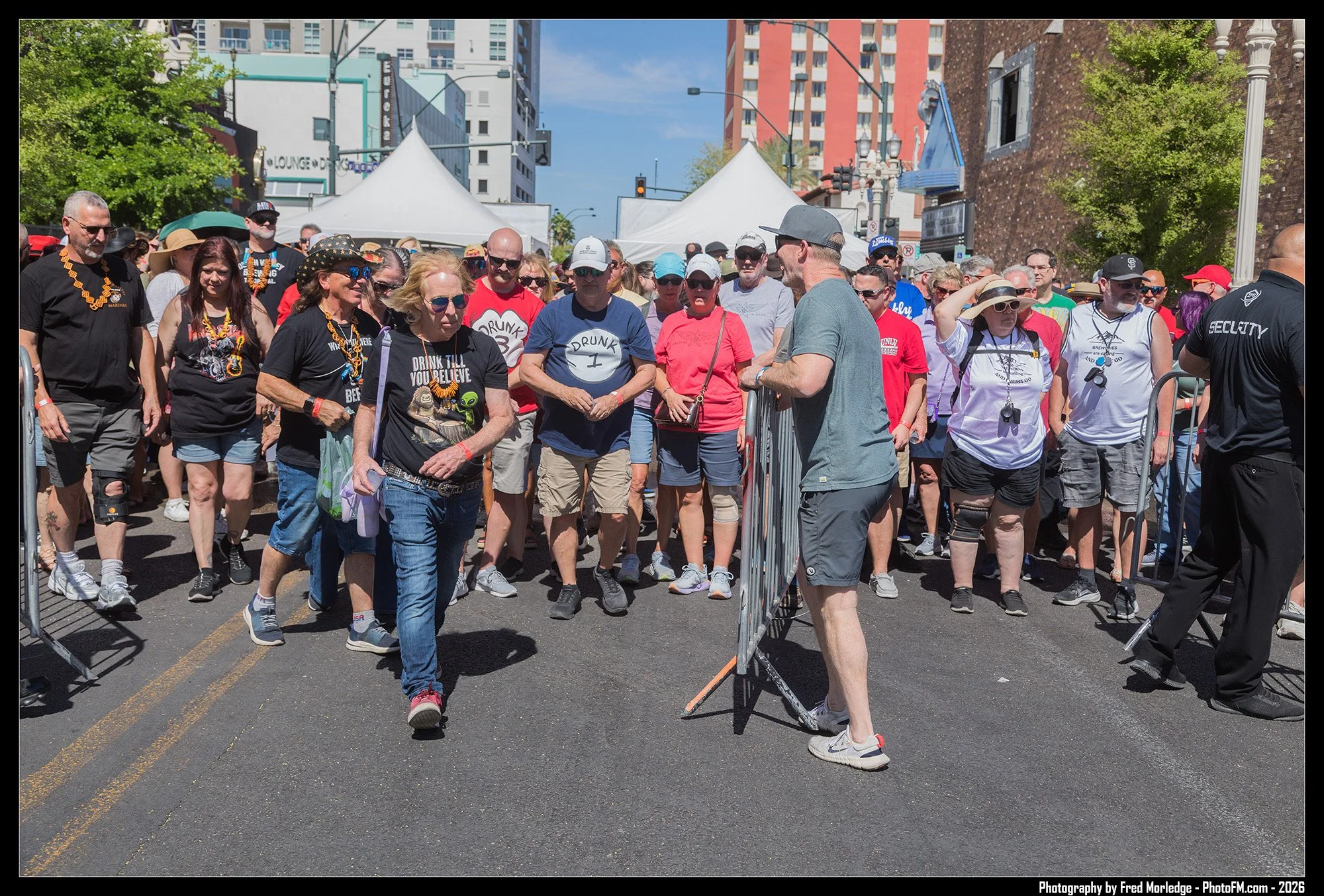 Crowd of people at an outdoor event in a city, waiting in front of a barrier with tents and buildings in the background on a sunny day.