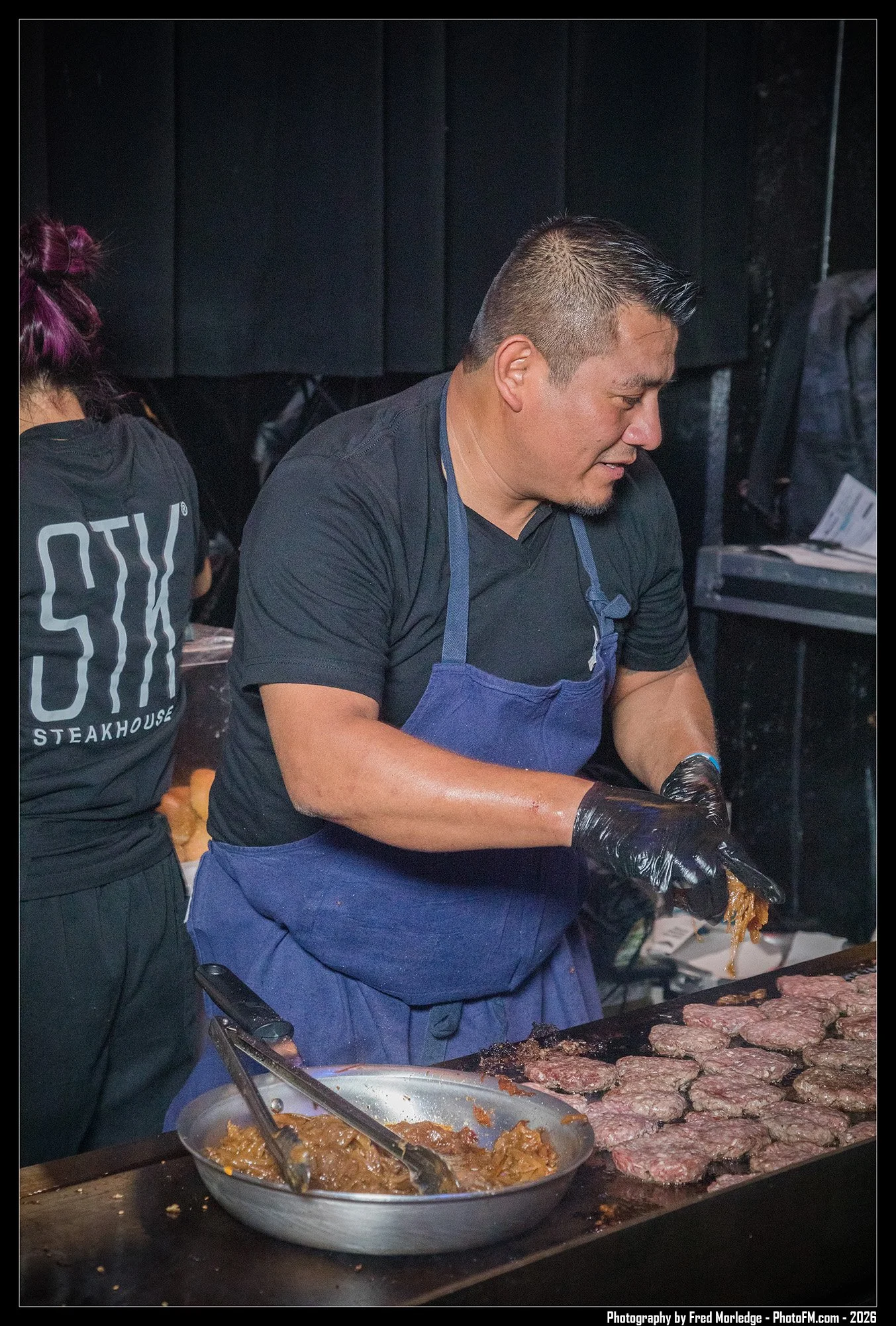A man wearing a black shirt and a blue apron cooks meat and other ingredients on a grill, with a bowl of sauce and a black glove on his right hand. Part of a woman with purple hair and a black T-shirt with white text is visible on the left.